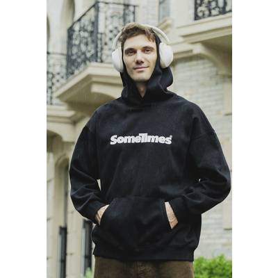 A man is wearing headphones and a black hoodie with the print 'Sometimes'. He is standing in front of a building with balconies.