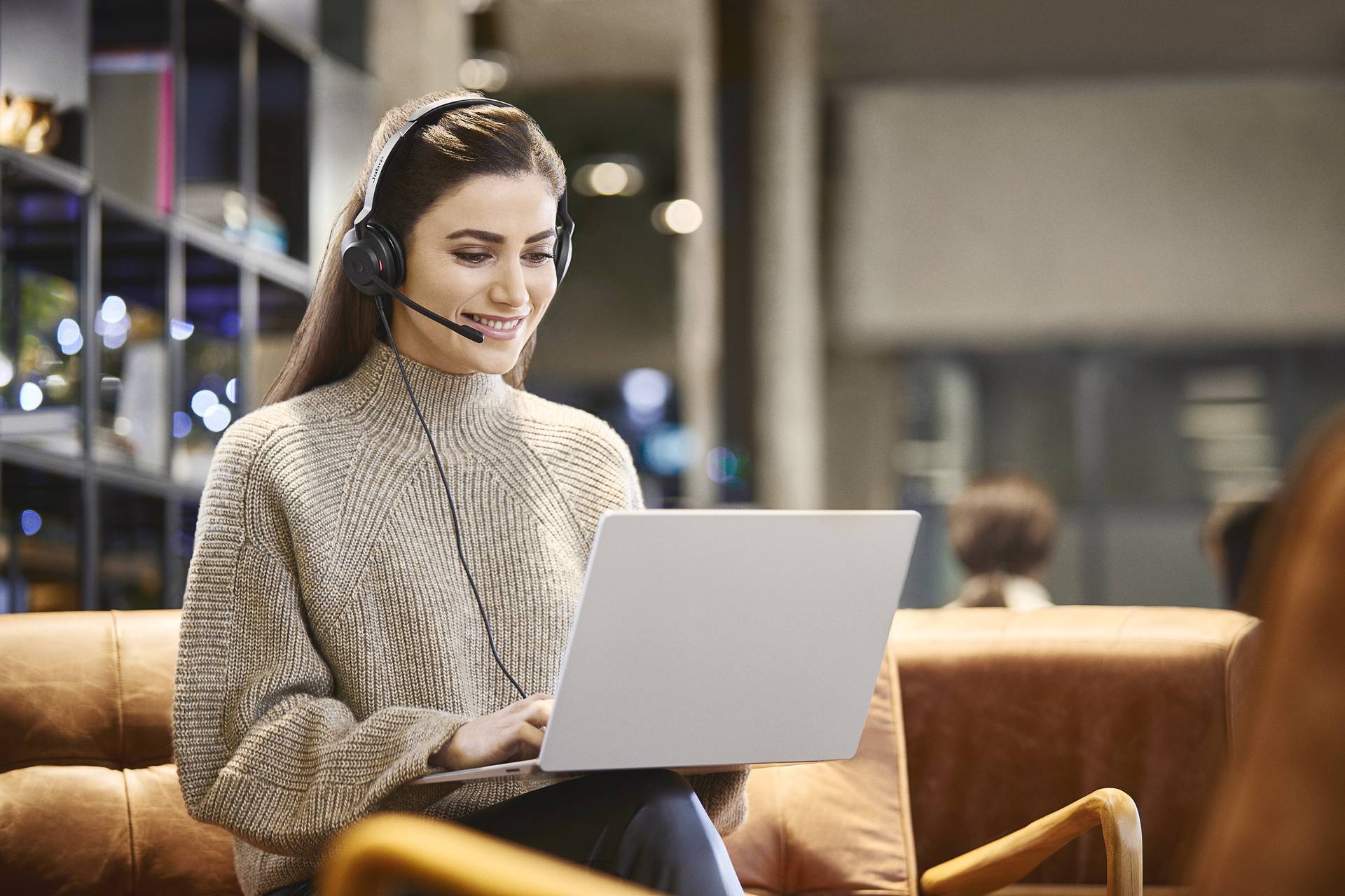 A woman wearing a headset sits in a modern office, smiling as she works on her laptop.
