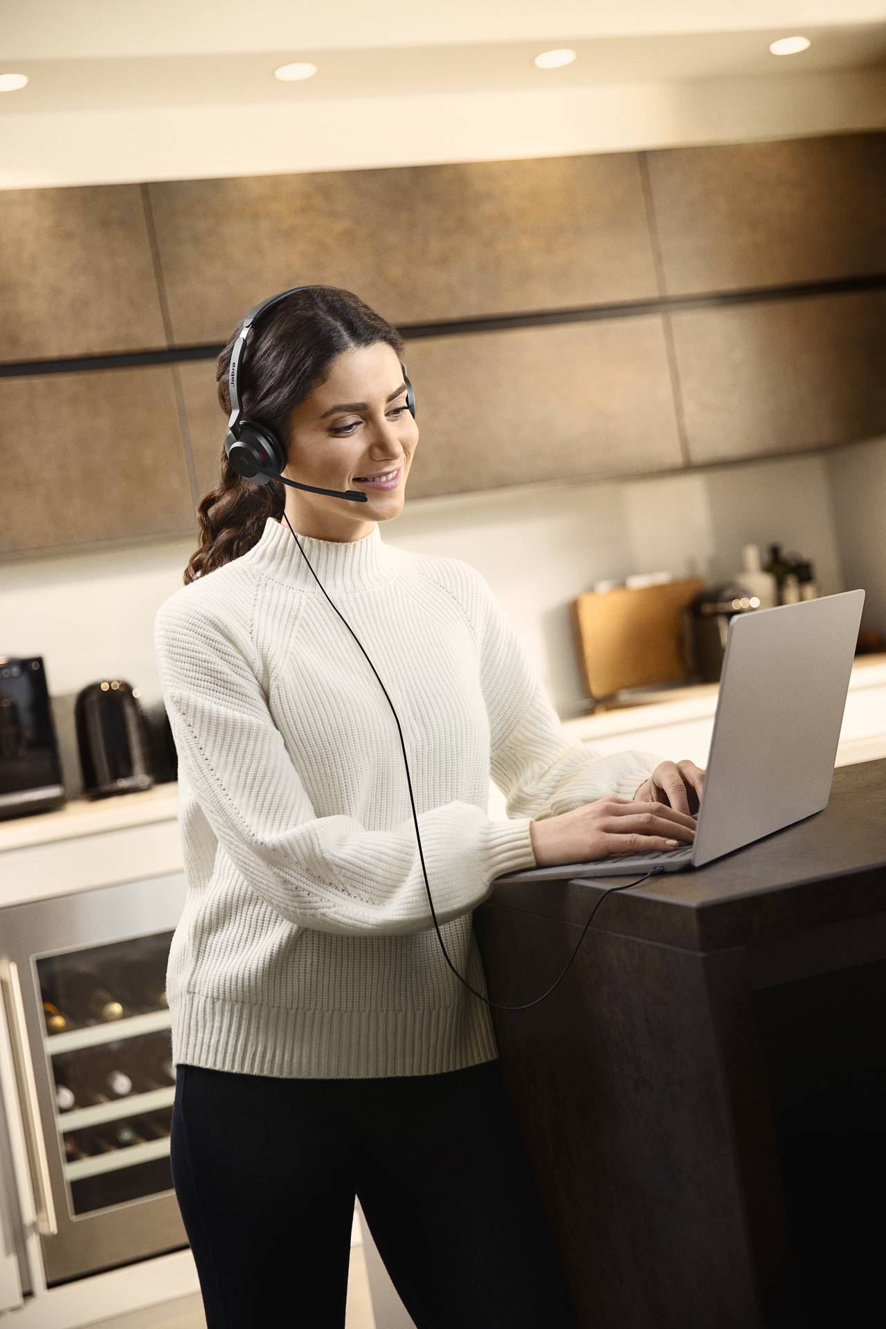 A woman wearing a headset is standing in a modern kitchen and typing on a laptop. She looks focused and is wearing a white jumper.