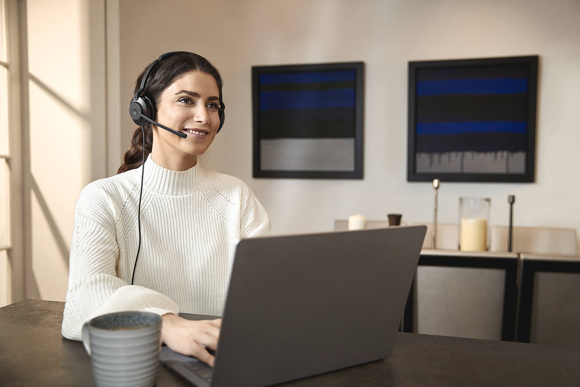 A woman sits smiling in front of a laptop with a headset, in a modern room. Abstract paintings are visible in the background.