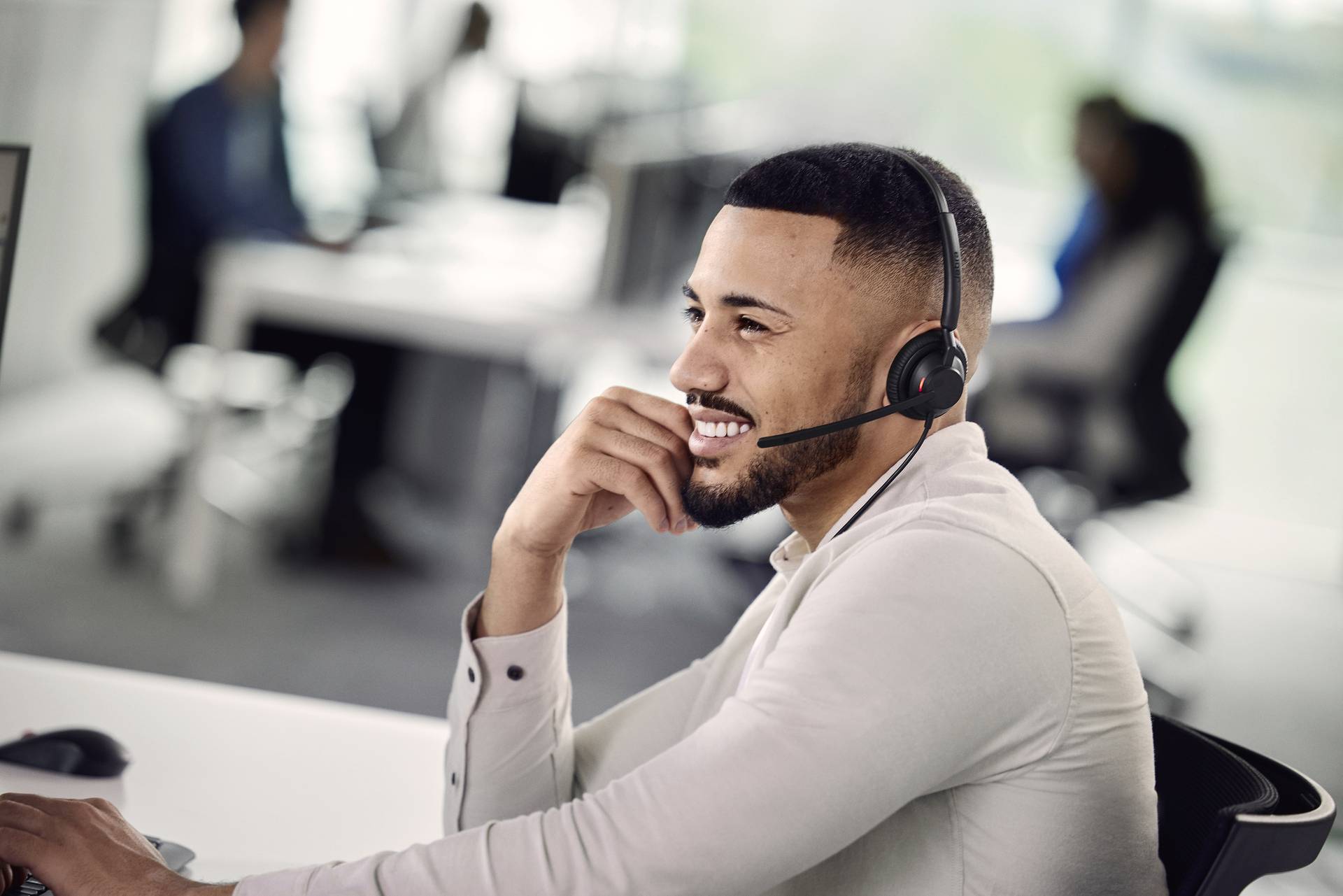 A man with a headset sits smiling at his desk in a modern office; customer service or call centre environment.