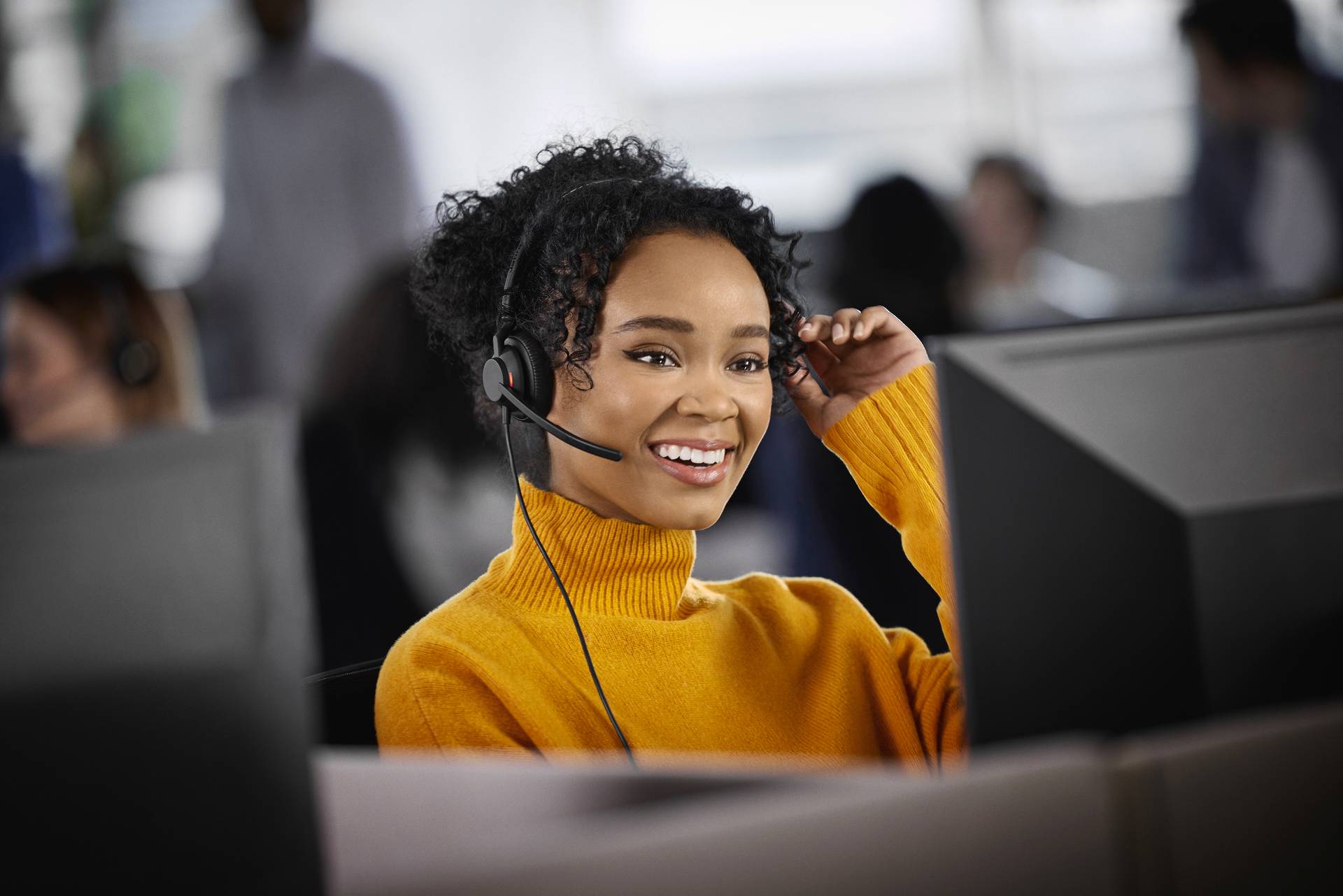 A person is sitting smiling in front of a computer and wearing a headset, possibly in a customer service office.