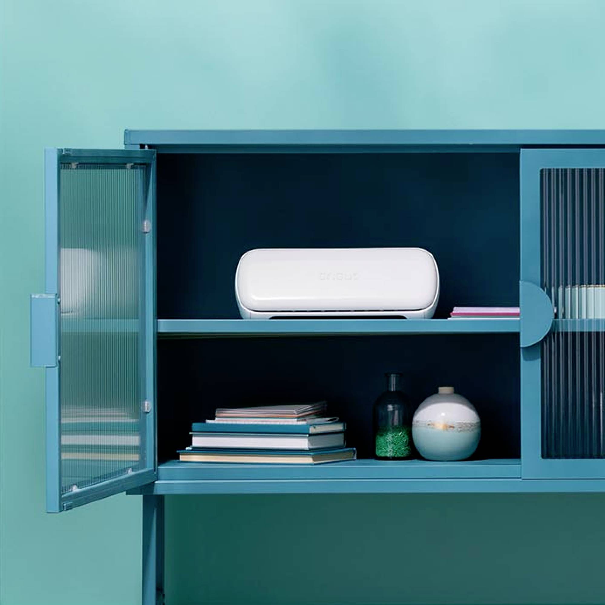 A blue cupboard with open doors reveals a white electronic device, books, and two bottles on two shelves.