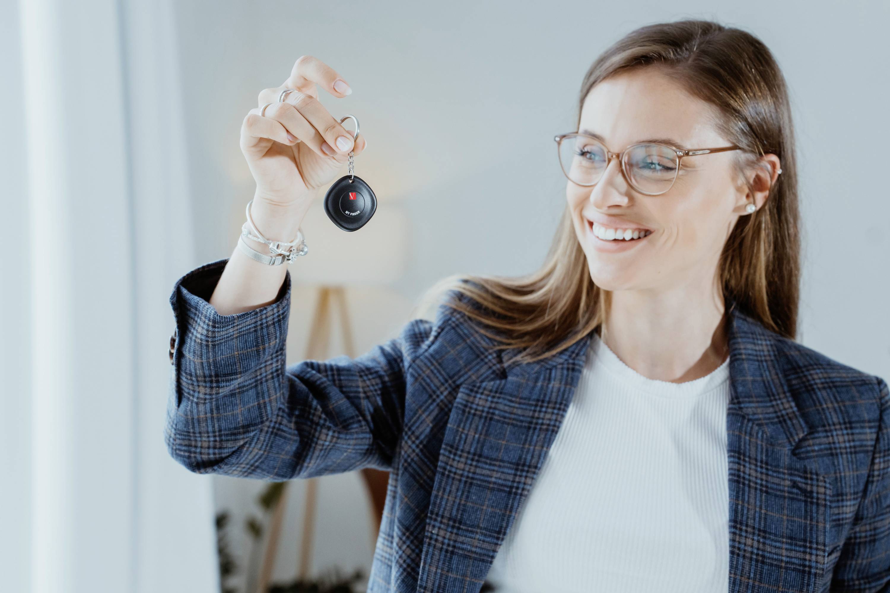 A smiling woman wearing glasses is holding a keyring up in the air. She is wearing a checked blazer and standing in front of a light background.