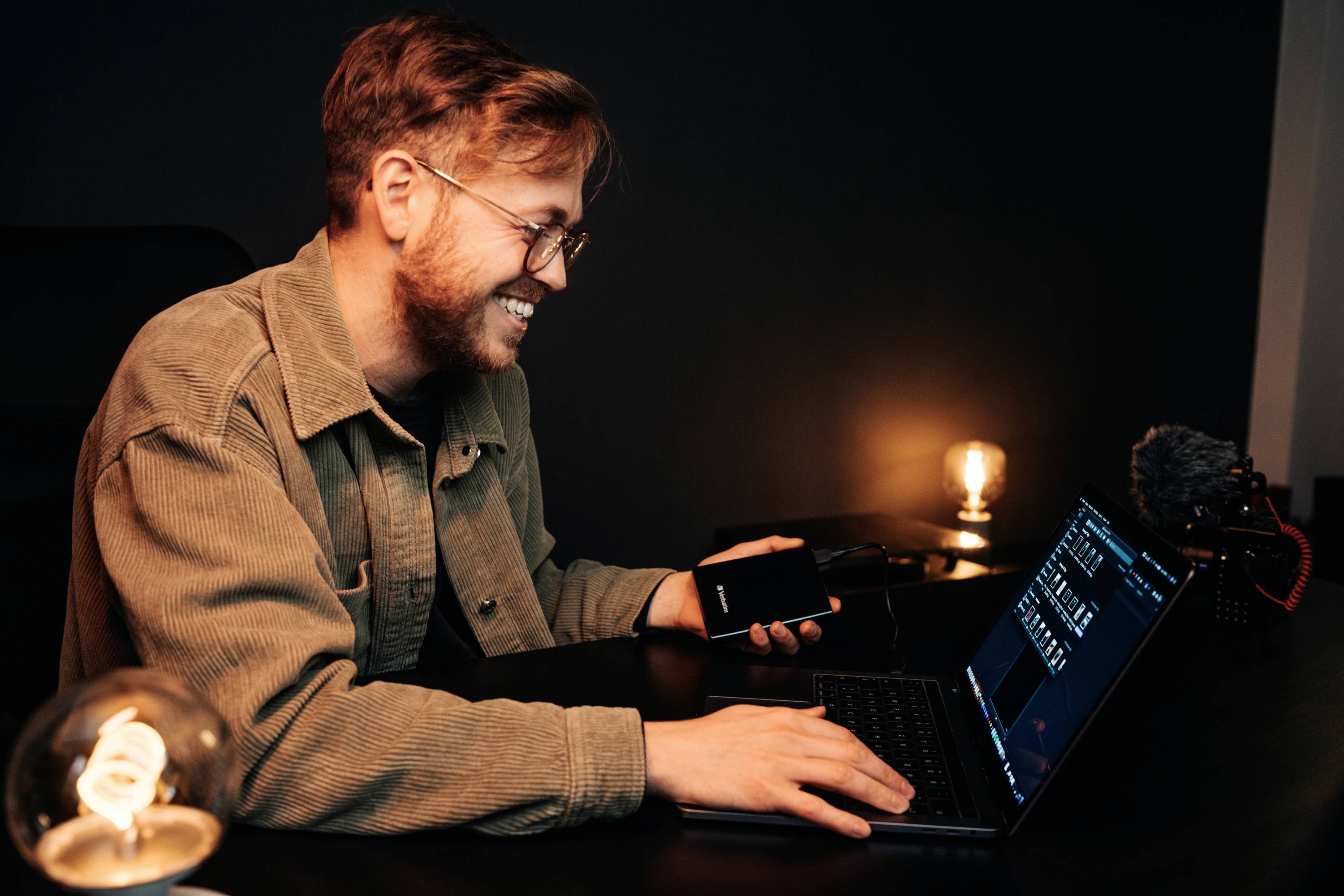 A man sits at a desk with a laptop and a mobile phone. In the background, a light bulb is glowing, creating a cosy atmosphere.