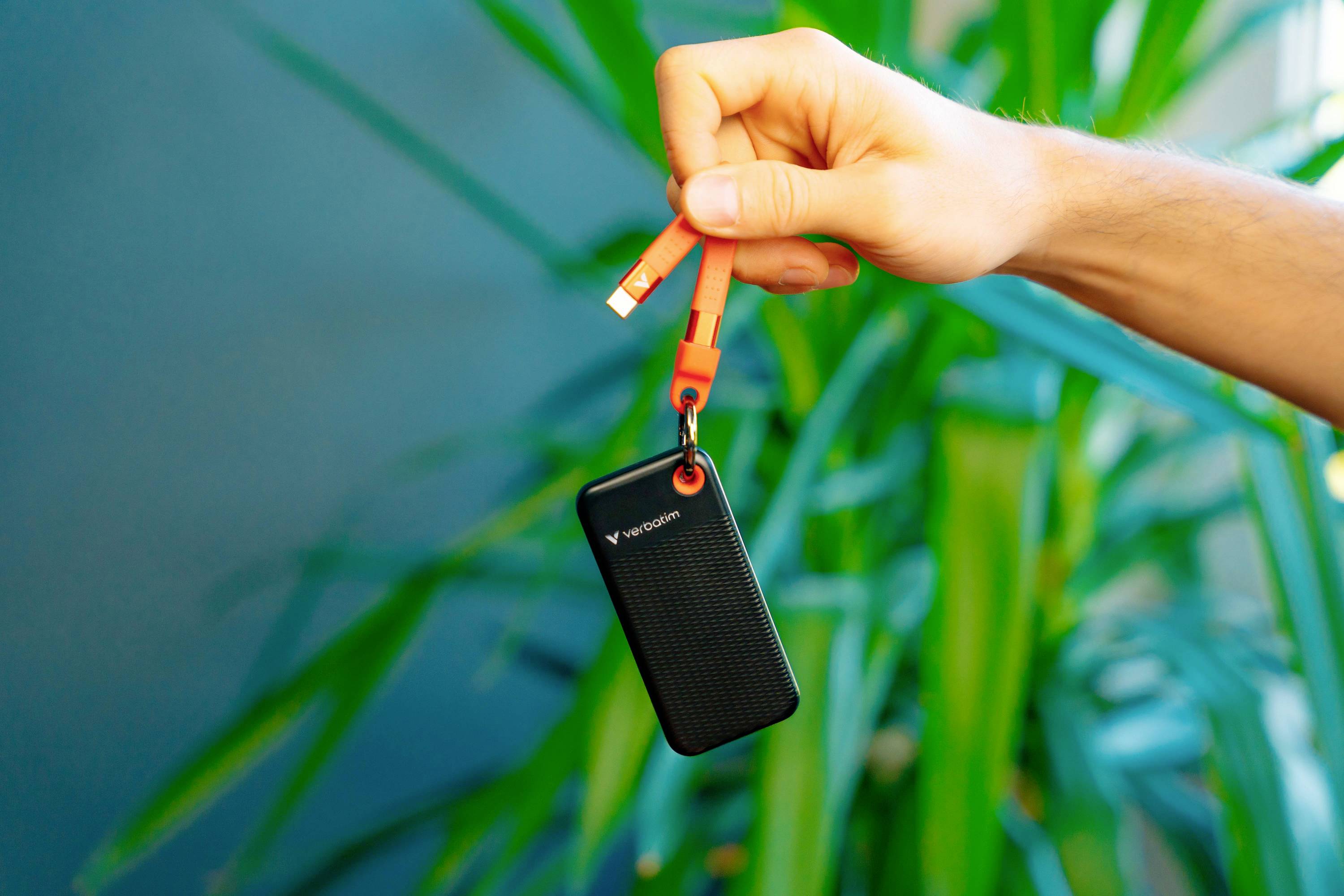A hand holds a small, black external storage device with an orange cable against a green plant background.