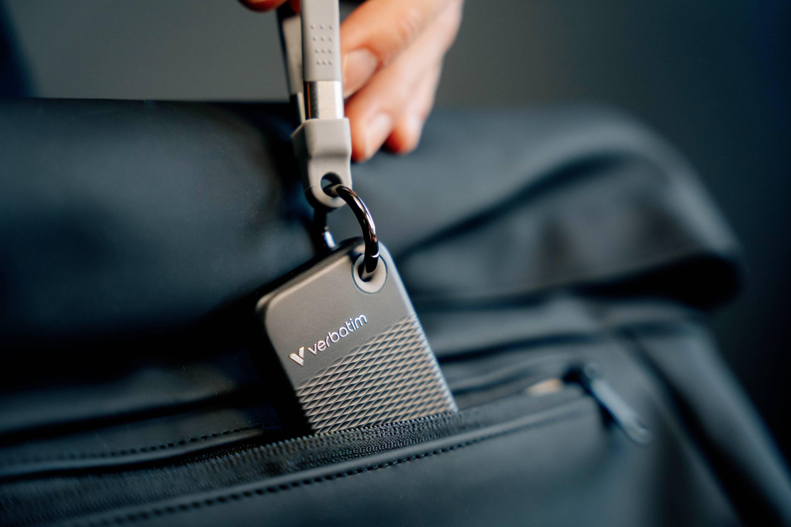 A hand is attaching a Verbatim USB stick to a black bag. The stick has a modern, structured design.