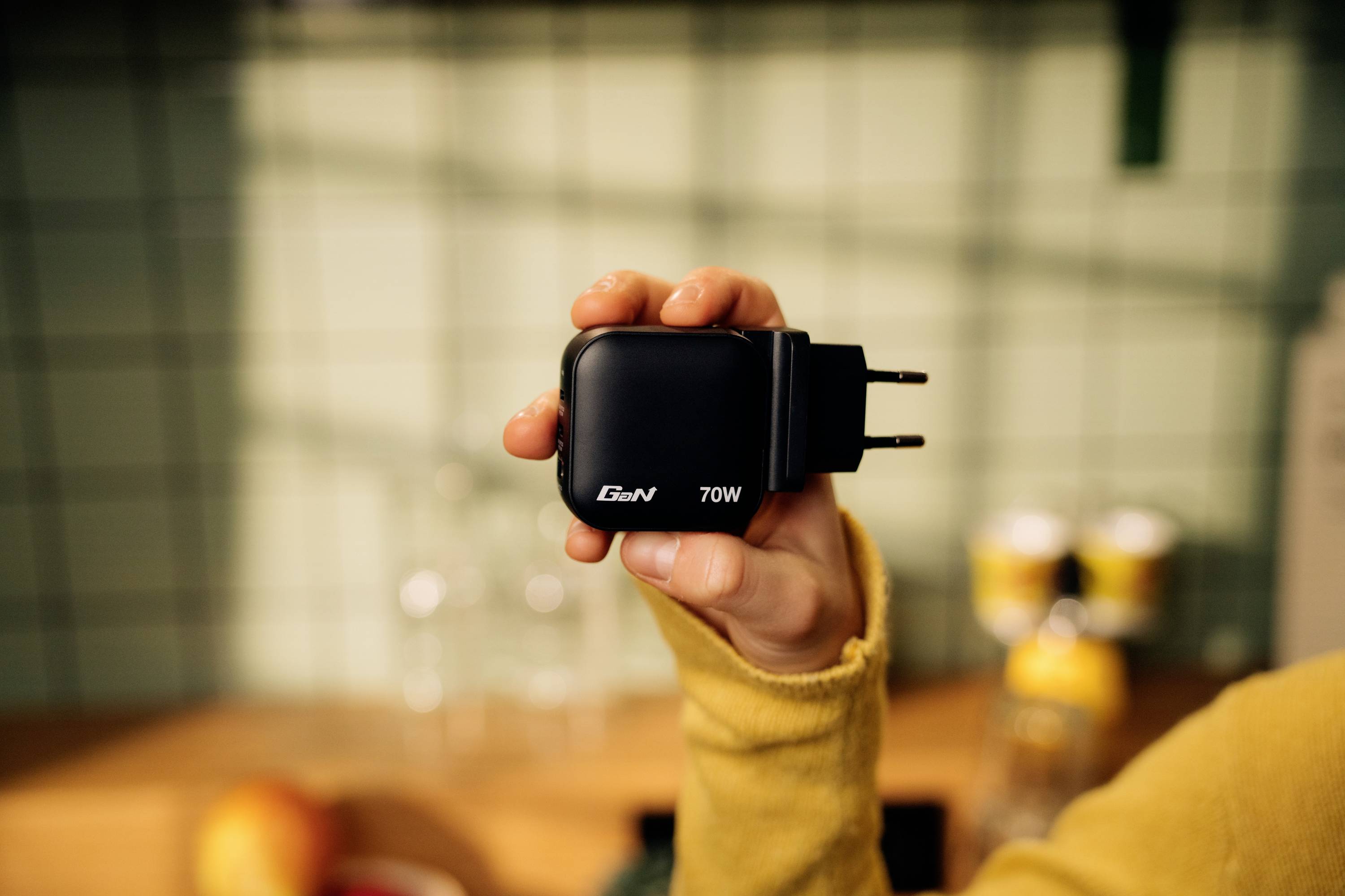 A hand is holding a black 70W charger with 'GaN' written on it against a blurred kitchen background.