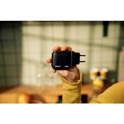 A hand is holding a black 70W charger with 'GaN' written on it against a blurred kitchen background.