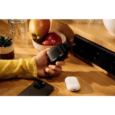 A hand is holding a black 70W charger on a wooden surface. Alongside it are a smartphone, wireless earbuds, and a bowl of apples.