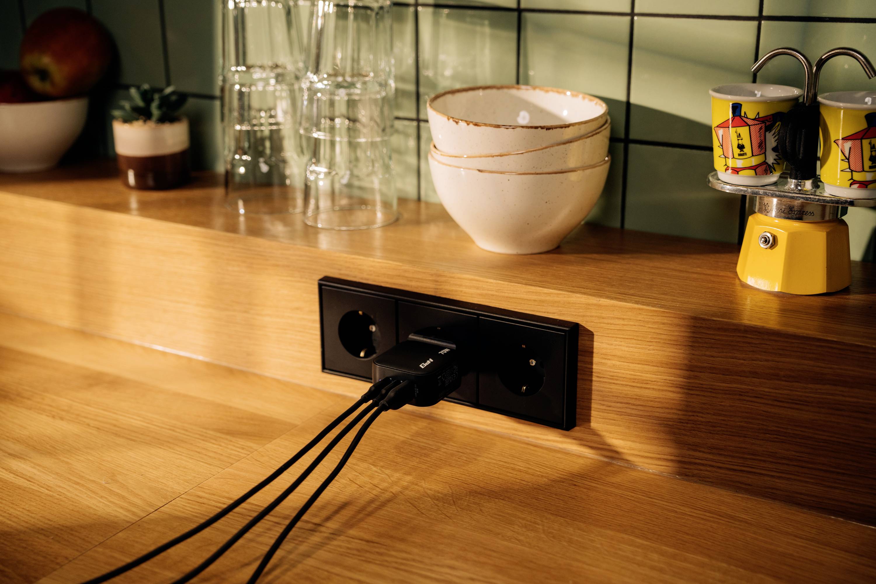 Three black cables are plugged into a socket on a wooden kitchen counter. Beside them are glasses, bowls, and cups.