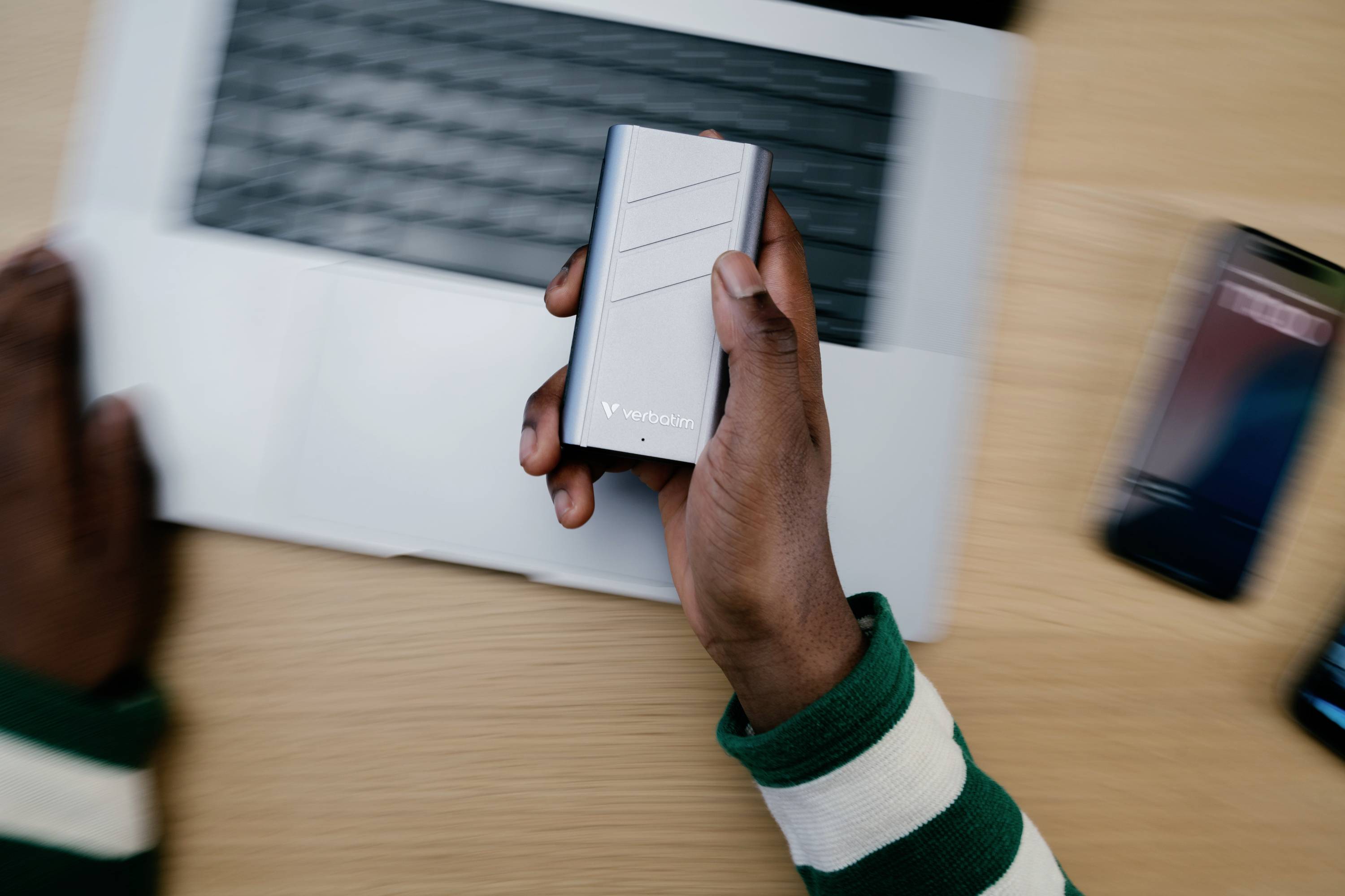 A person is holding a portable hard drive in front of a laptop. The notebook is open, and a smartphone is lying on the table beside it.