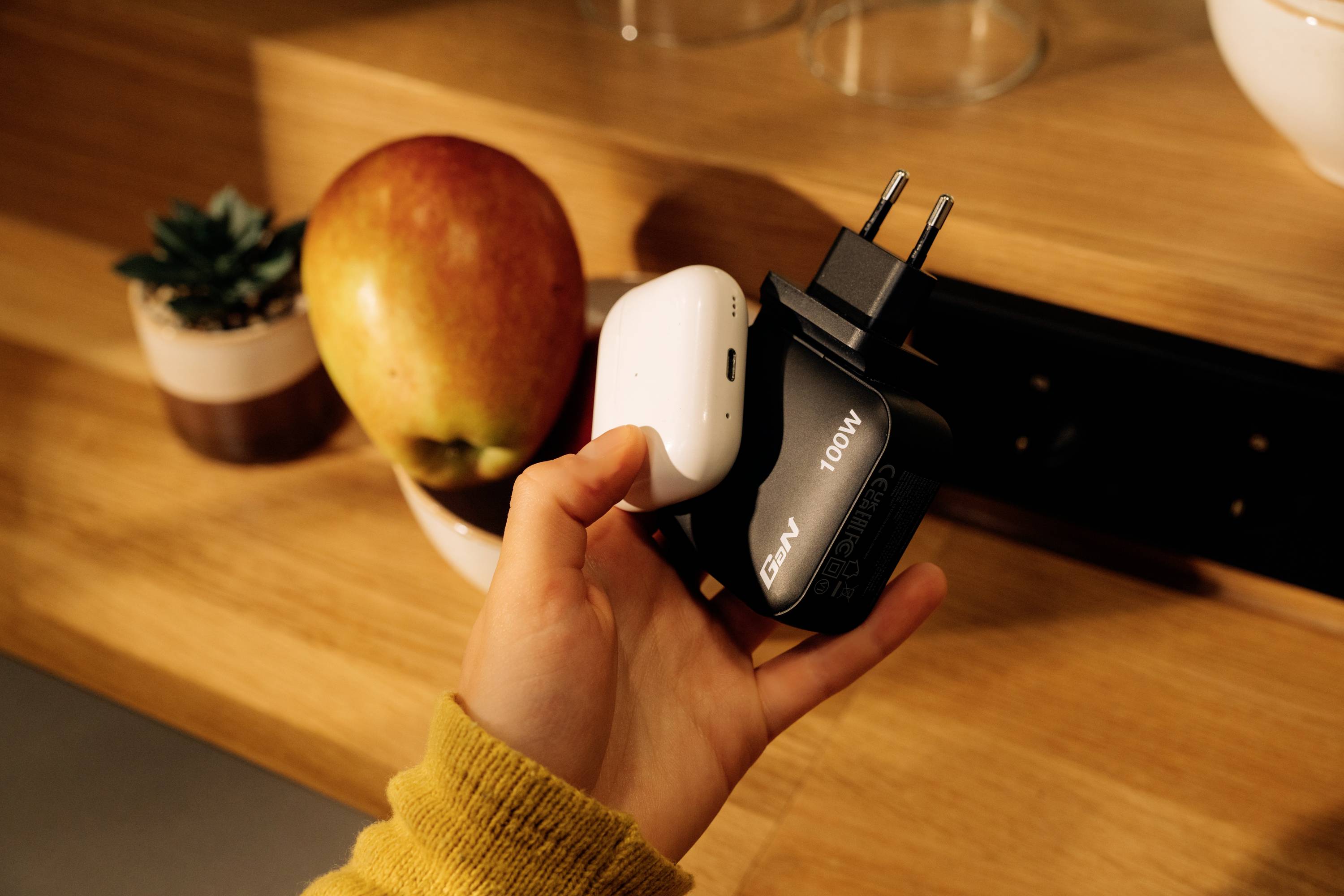 A hand is holding a white and a black charger next to a table with an apple and a potted plant in a well-lit room.
