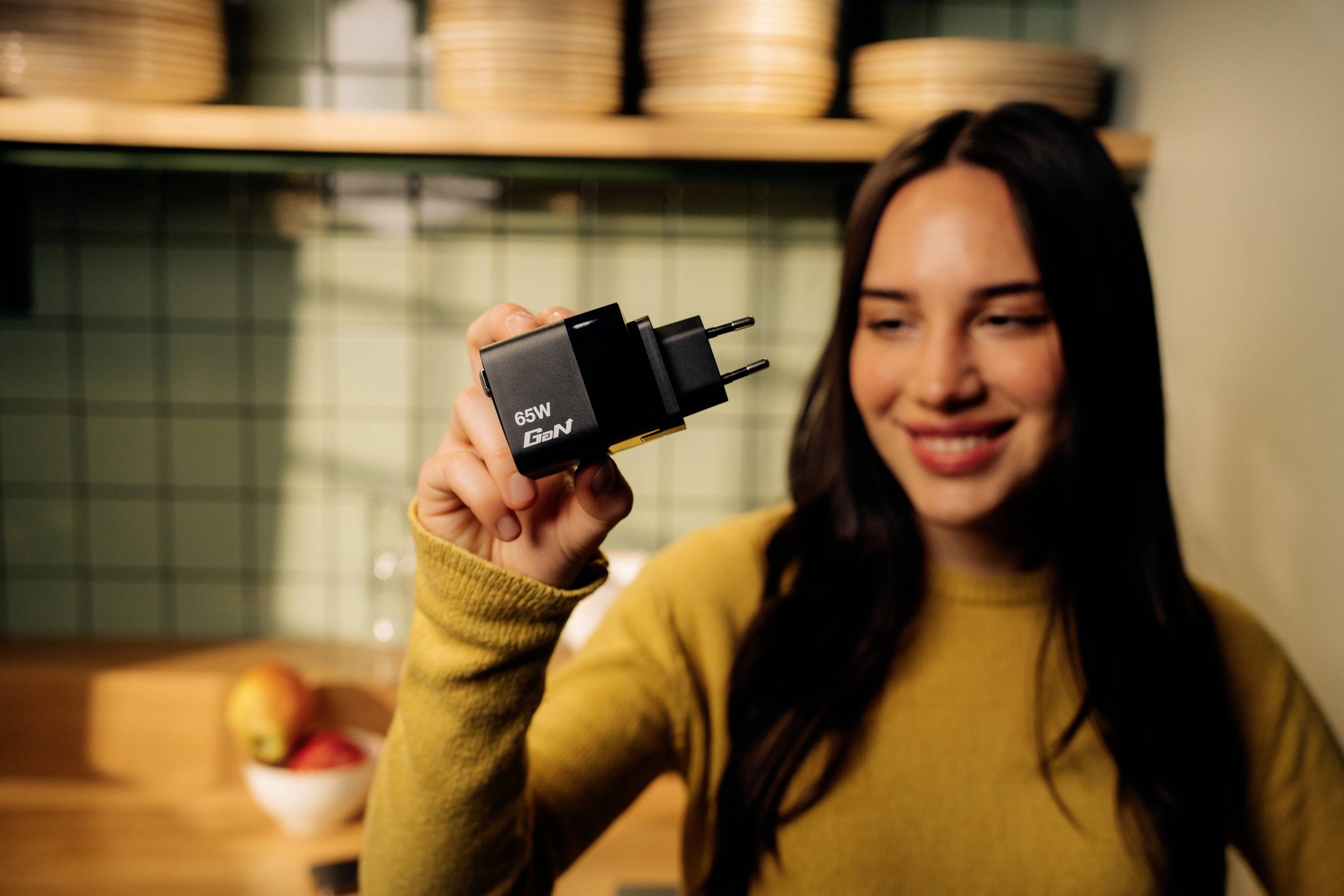 Woman in a yellow jumper holding a black charger with '65W GaN' printed on it. Background shows a kitchen.