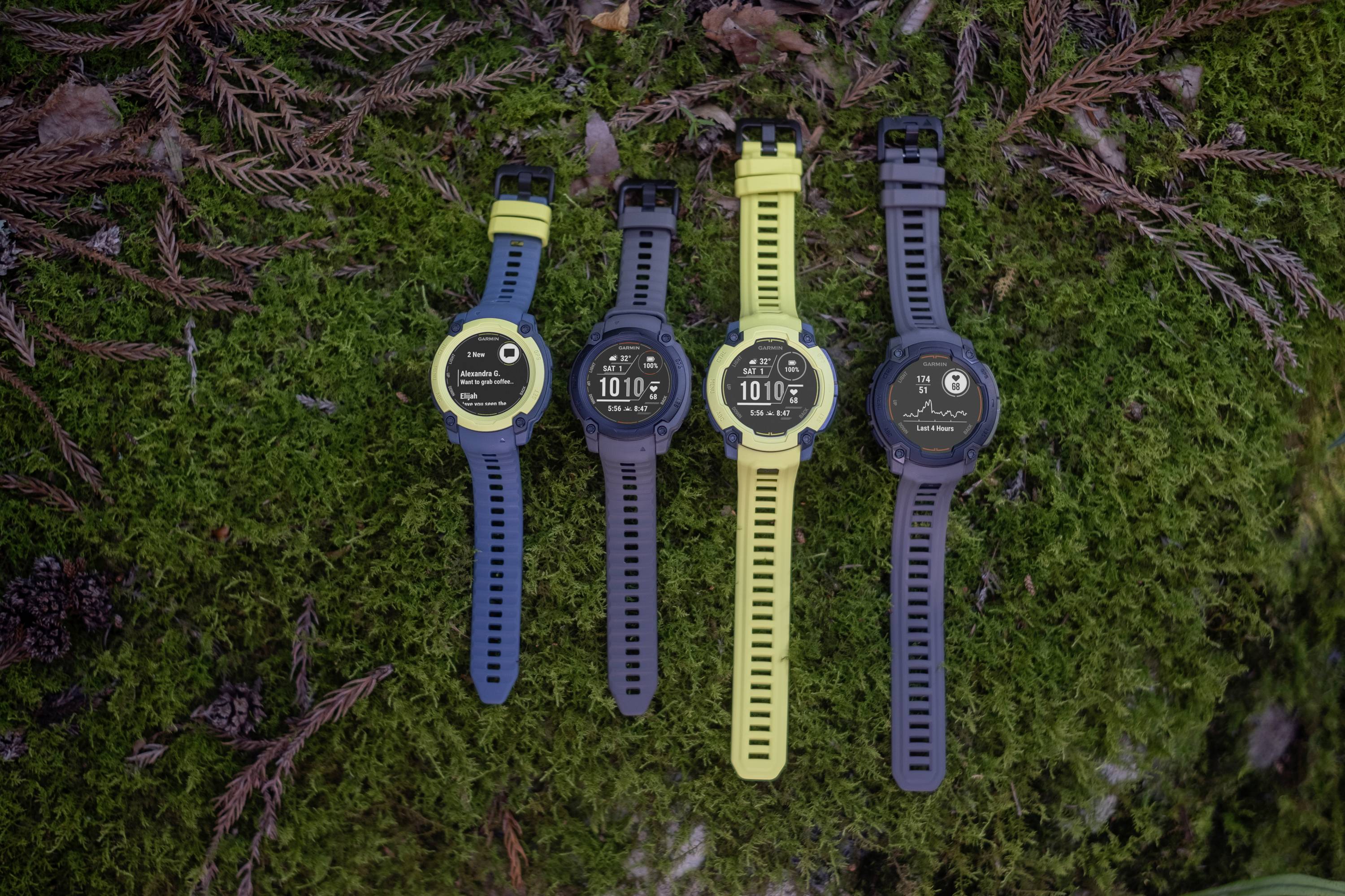 Four colourful outdoor smartwatches on moss-covered ground, each displaying different screens.