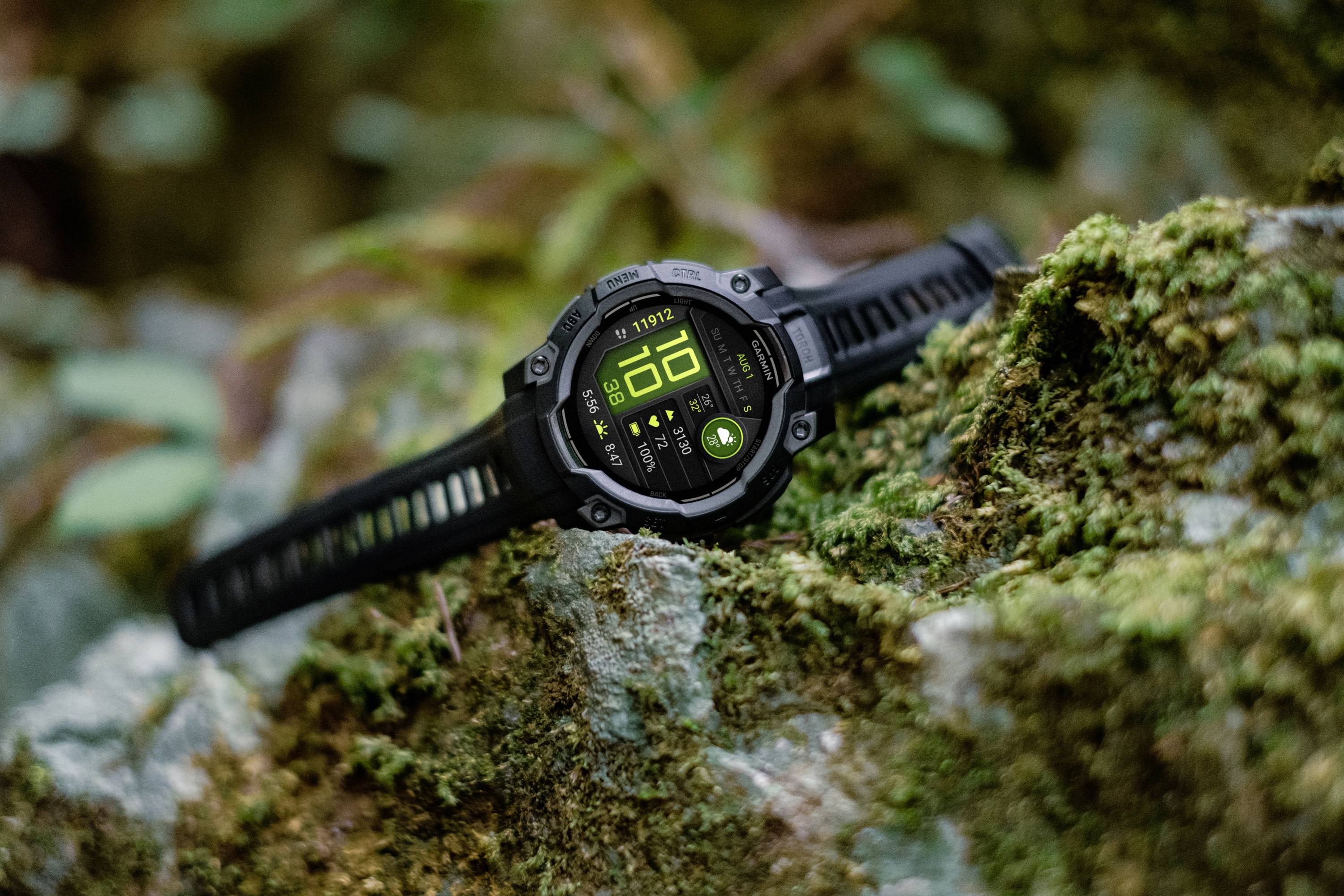 A black outdoor smartwatch with a digital display is resting on a moss-covered rock. Plants can be seen in the background.