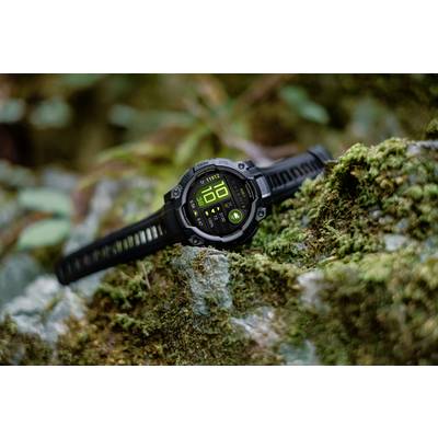 A black outdoor smartwatch with a digital display is resting on a moss-covered rock. Plants can be seen in the background.