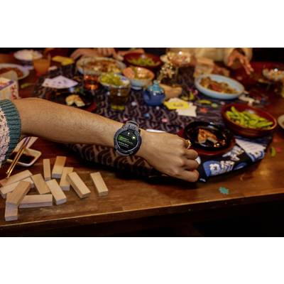 A person displays a smartwatch on their wrist. In the background, a table with food, drinks, and a Jenga game.