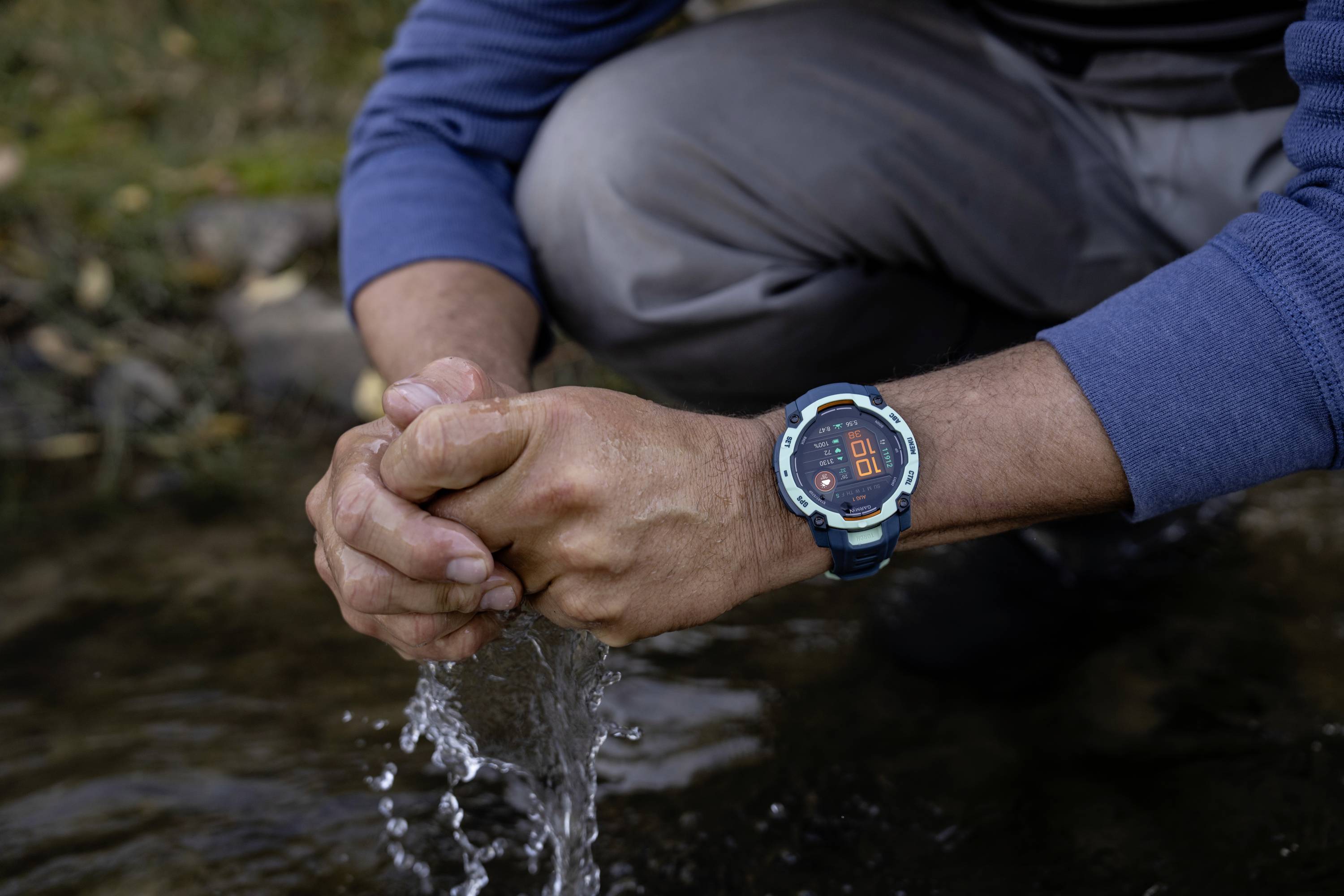 A person kneels by a river and collects water with their hands. They are wearing a wristwatch with a red display.
