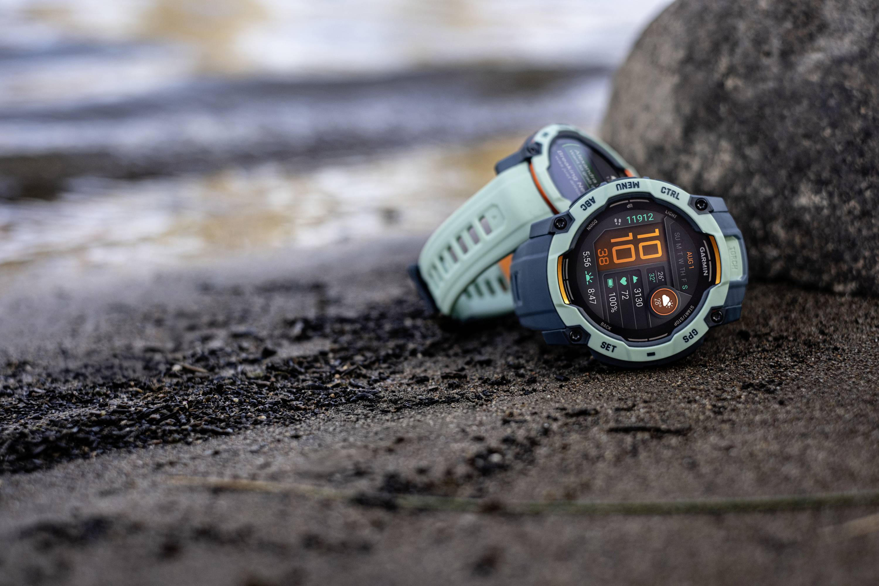 Two sporty wristwatches are lying on a rocky beach. The dial of one watch shows the numeral '10'. Water is visible in the background.