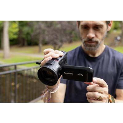 A man is holding a Panasonic 4K video camera outdoors and carefully checking the lens. Trees are visible in the background.