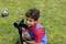 A boy sits on a grass verge and films with a video camera mounted on a tripod. A football lies on the grass in the background.