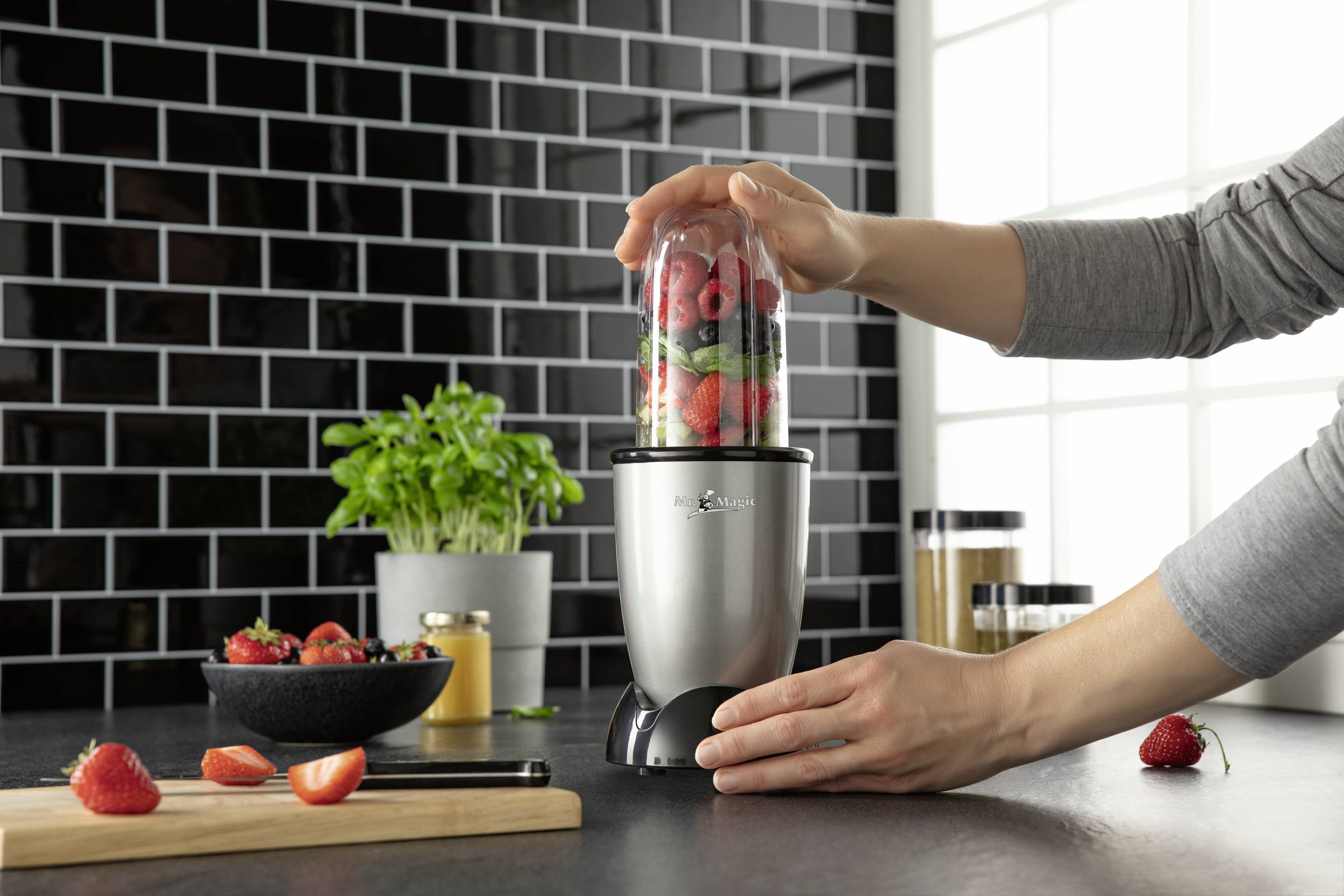 A person is using a blender to puree fruits. On the worktop, there is a bowl of berries and a wooden chopping board with half a strawberry.