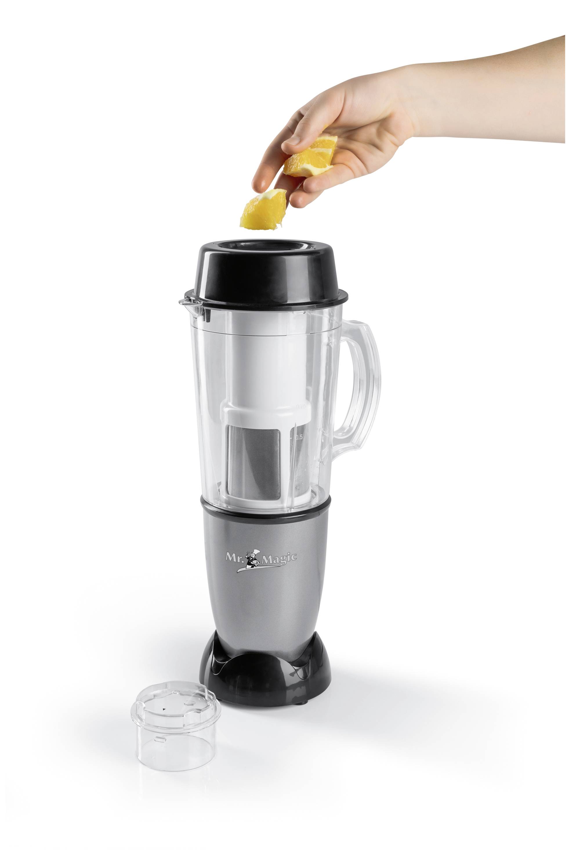 A hand places a piece of fruit into a blender. The blender has a silver base and a transparent container.