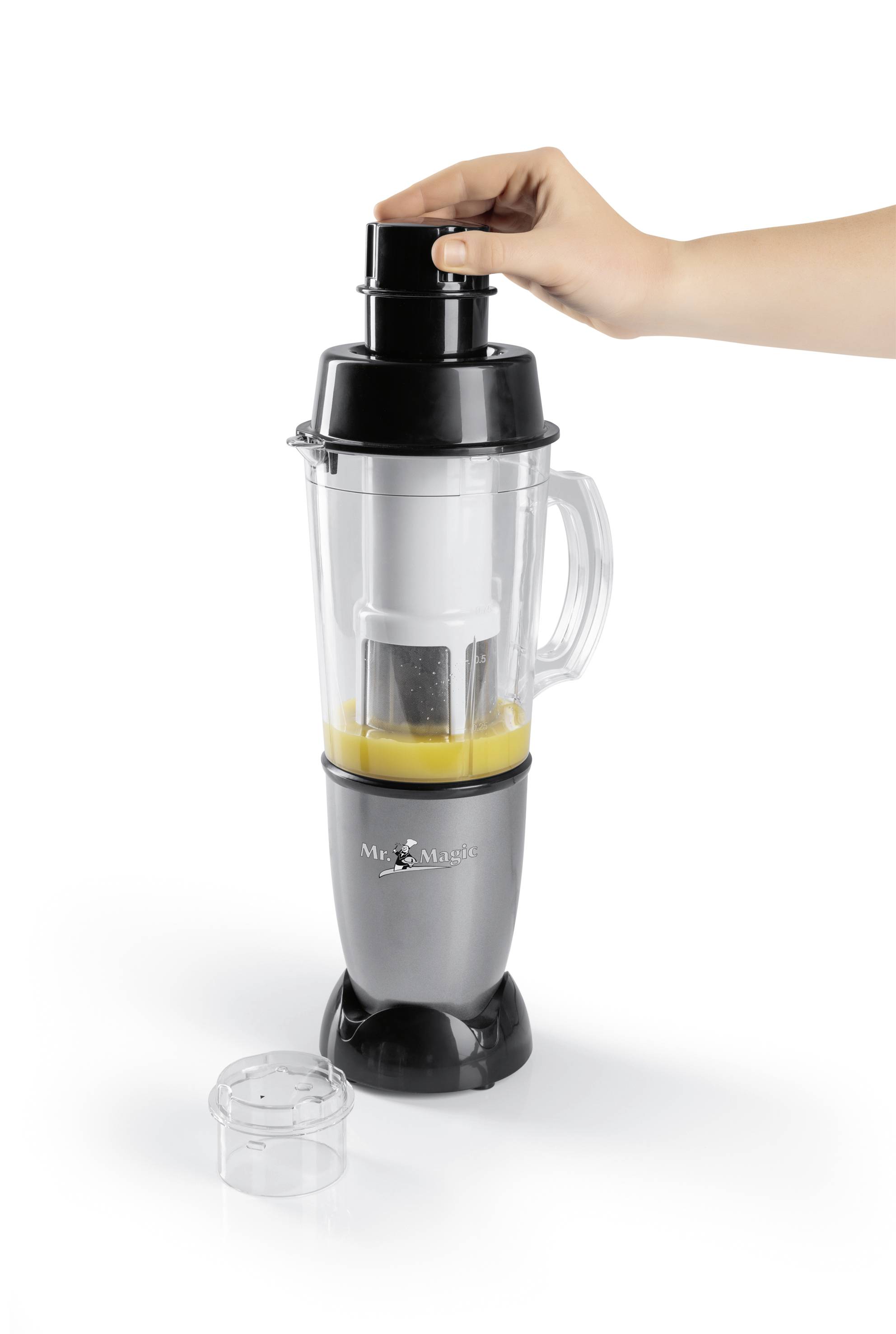 A hand is operating a blender containing yellow liquid in the container. A removable lid is lying beside it.