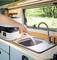 A person is filling water into a sink in a motorhome. A compact device with a hose is situated on the worktop.