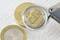 A magnifying glass focuses on a coin embossed with 'CEN.' Another coin lies beside it. The scene suggests examination of currency.