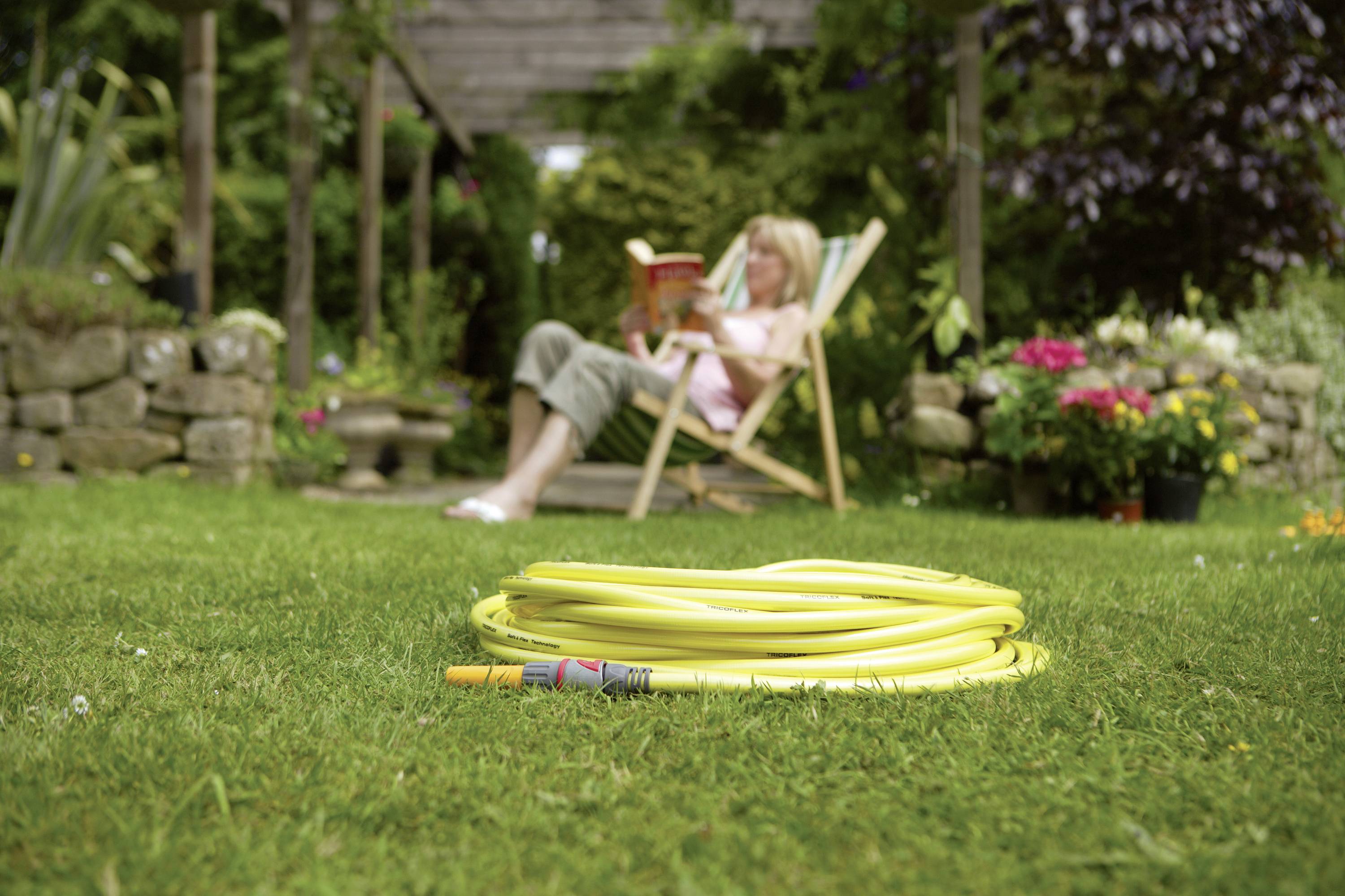 A yellow garden hose reel lies in the foreground on the lawn. In the background, a person sits on a deckchair reading a book.