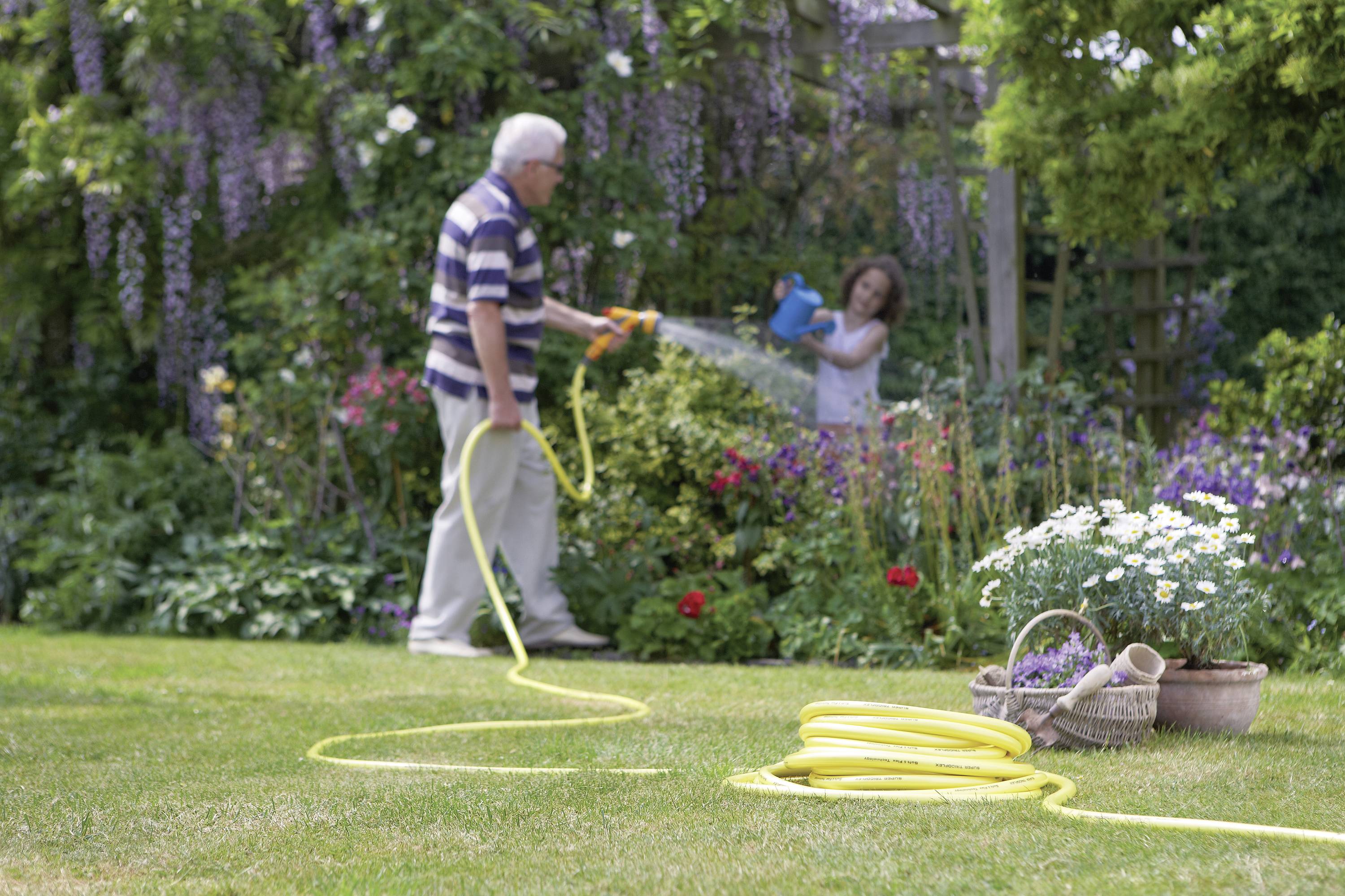 An older man is watering the garden with a hosepipe, while a girl waters plants with a watering can; a blooming garden in the background.