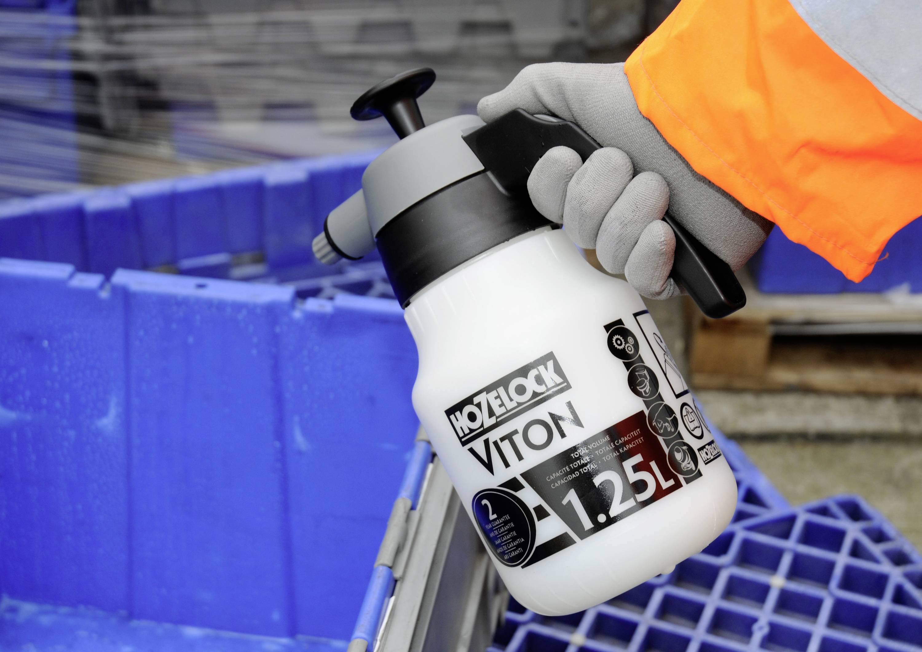 A hand wearing work gloves is holding a white spray container labelled 'Hozelock Viton 1.25L' in front of a blue pallet.