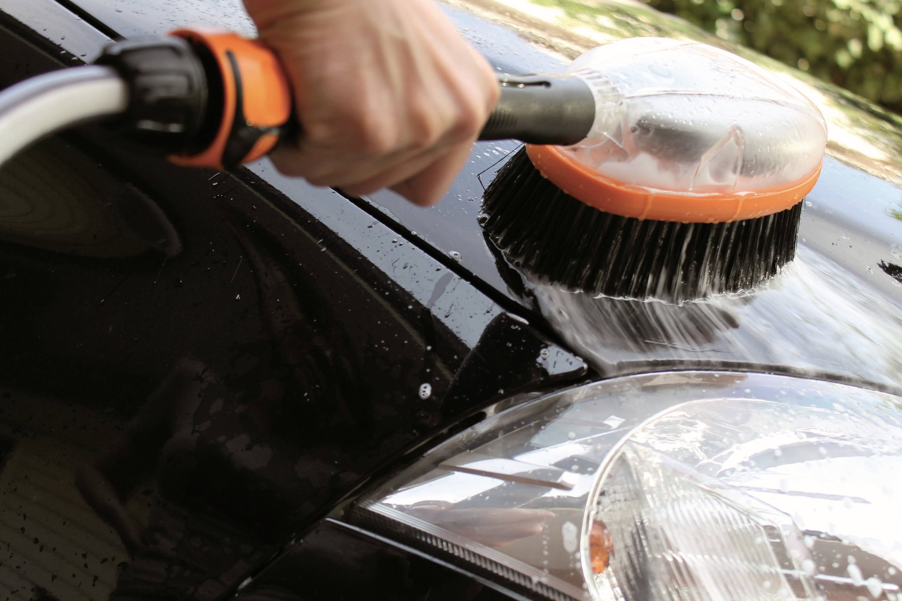 'Close-up of a hand cleaning a wet car with a brush. The focus is on the vehicle's front and headlight.'