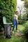 A person is raking leaves in a garden into a large waste bin. Surrounded by trees and shrubs, they are wearing casual clothing.