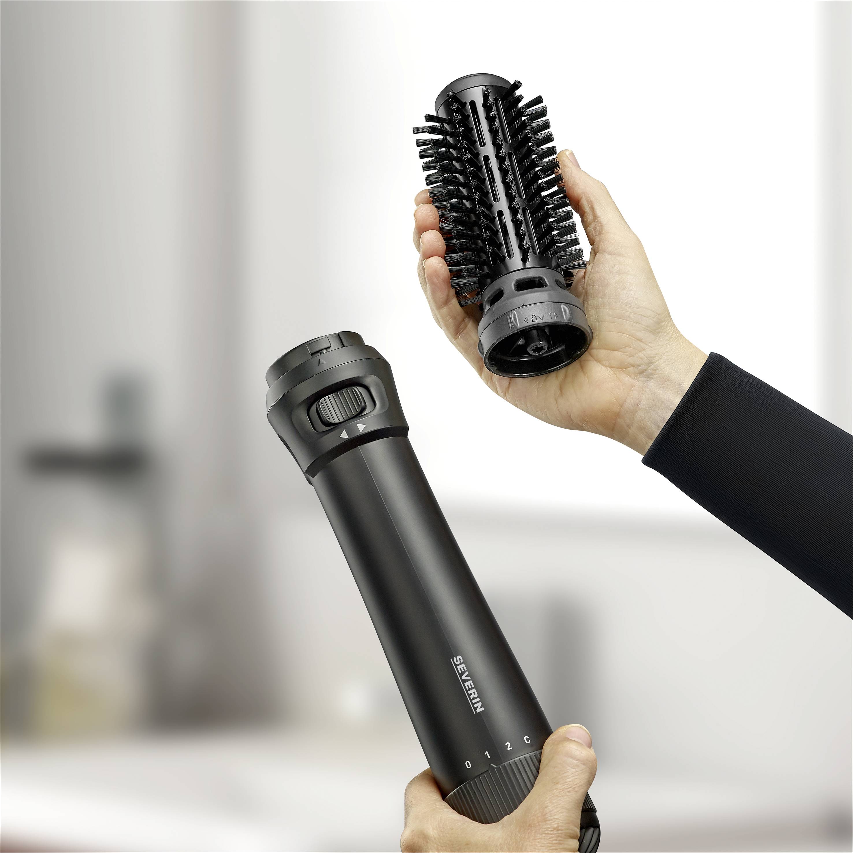 A person is holding a round hairbrush attachment attached to a black hair styler. In the background, a blurred room is visible.