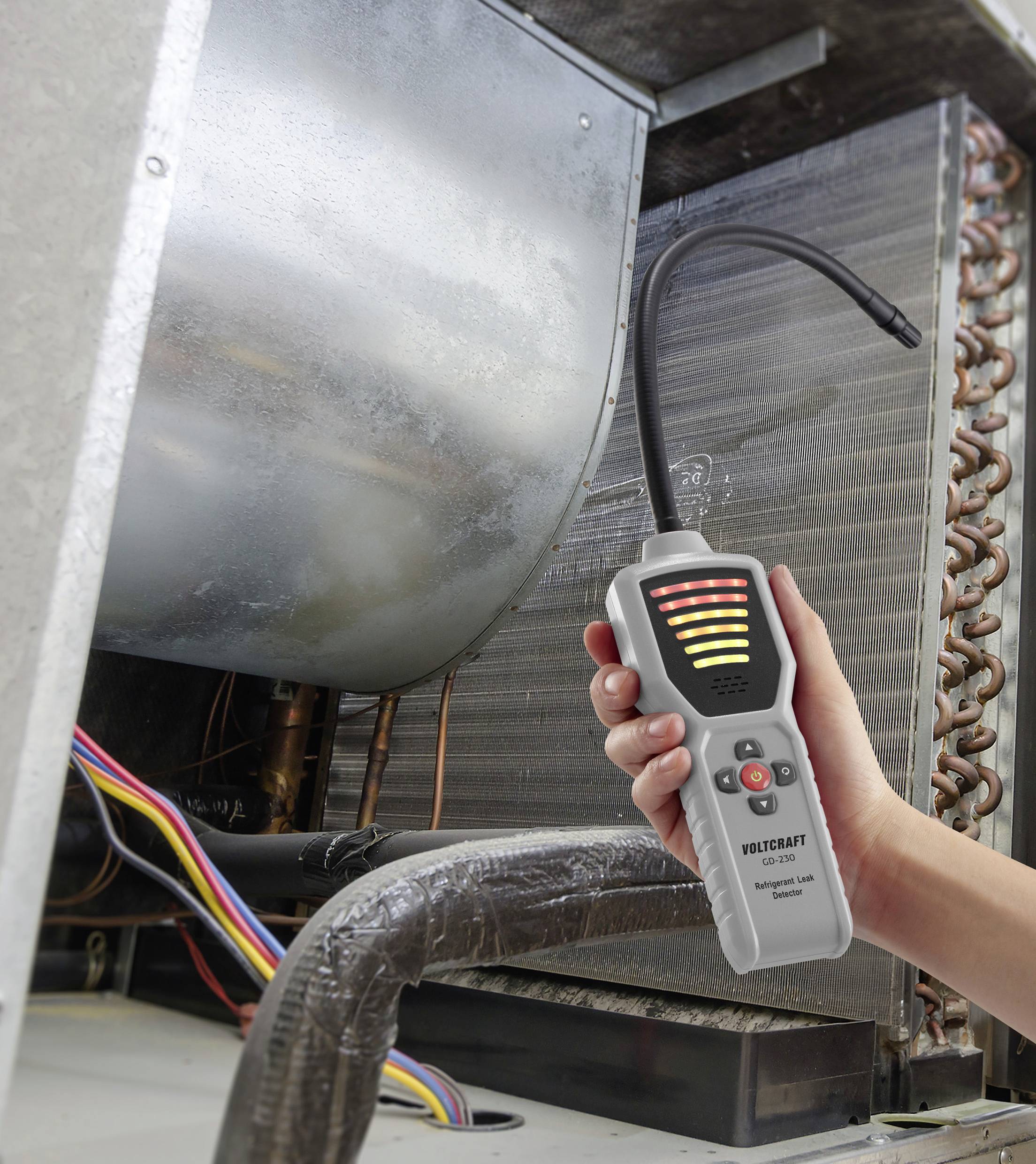 A hand holds an electronic leak detector with a flexible probe near HVAC equipment, displaying detection lights and control buttons.