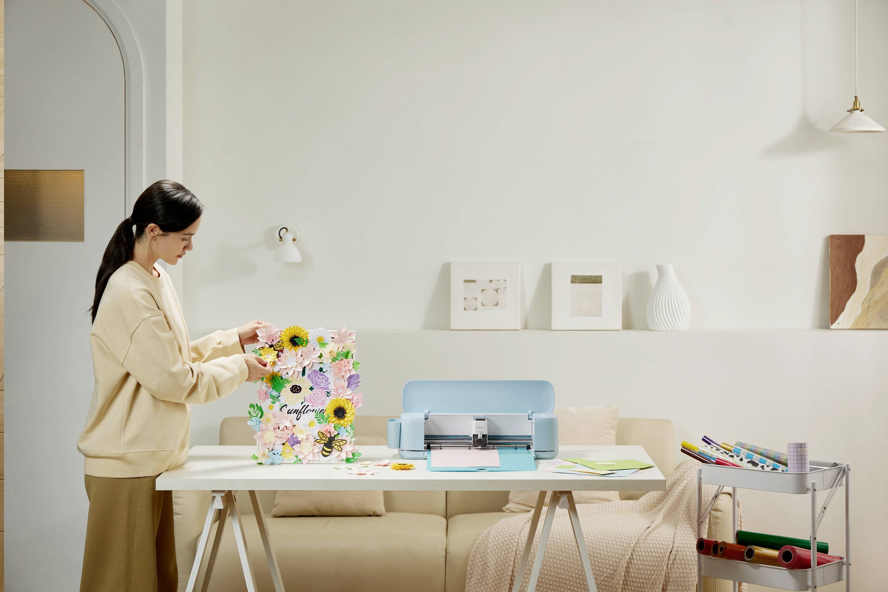 A woman is standing at a table, working on a colourful flower project with a cutting machine in a bright room.