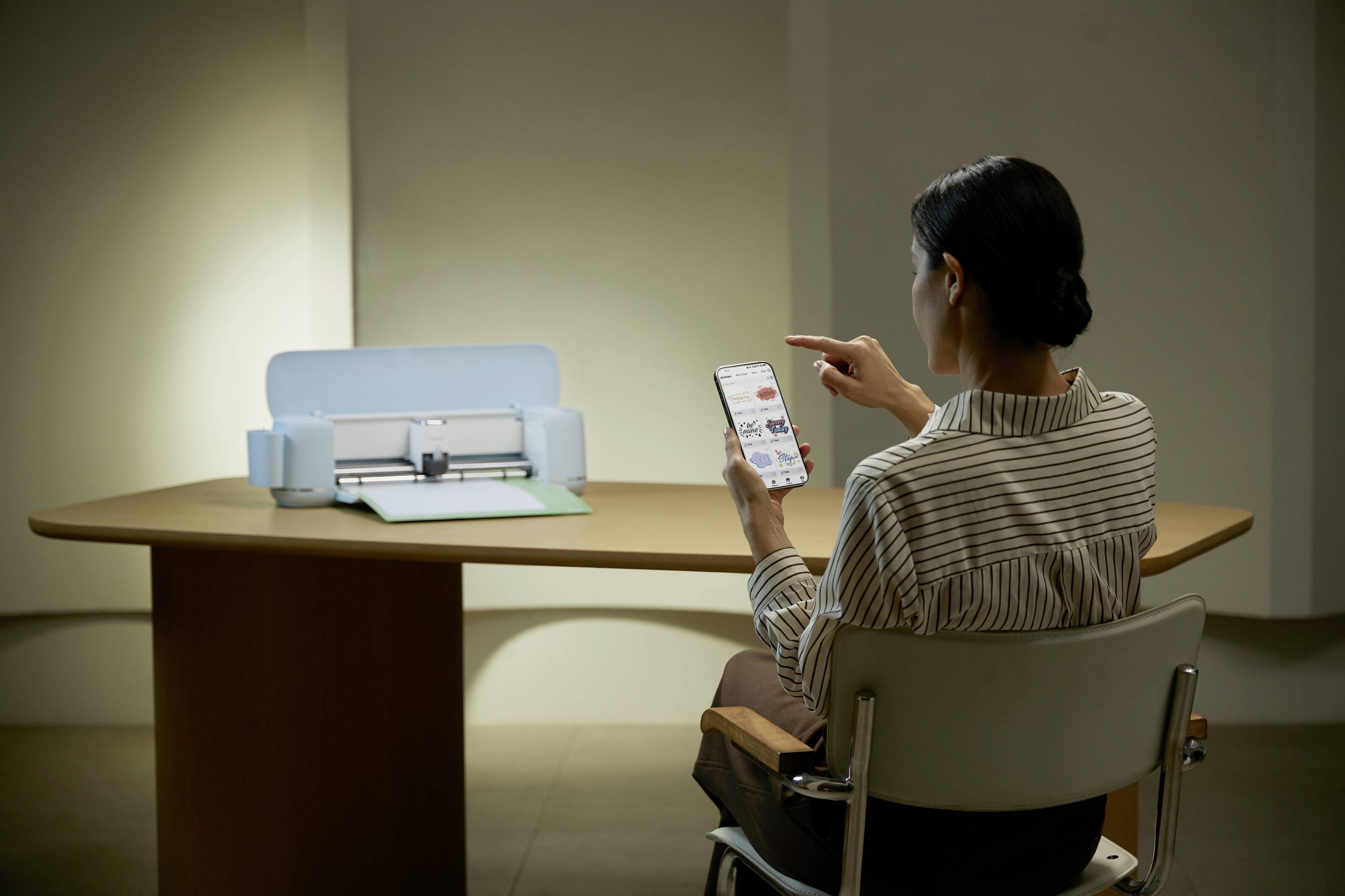 A woman is sitting at a table and using a smartphone, while a blue device, presumably a cutting plotter, sits on the table.