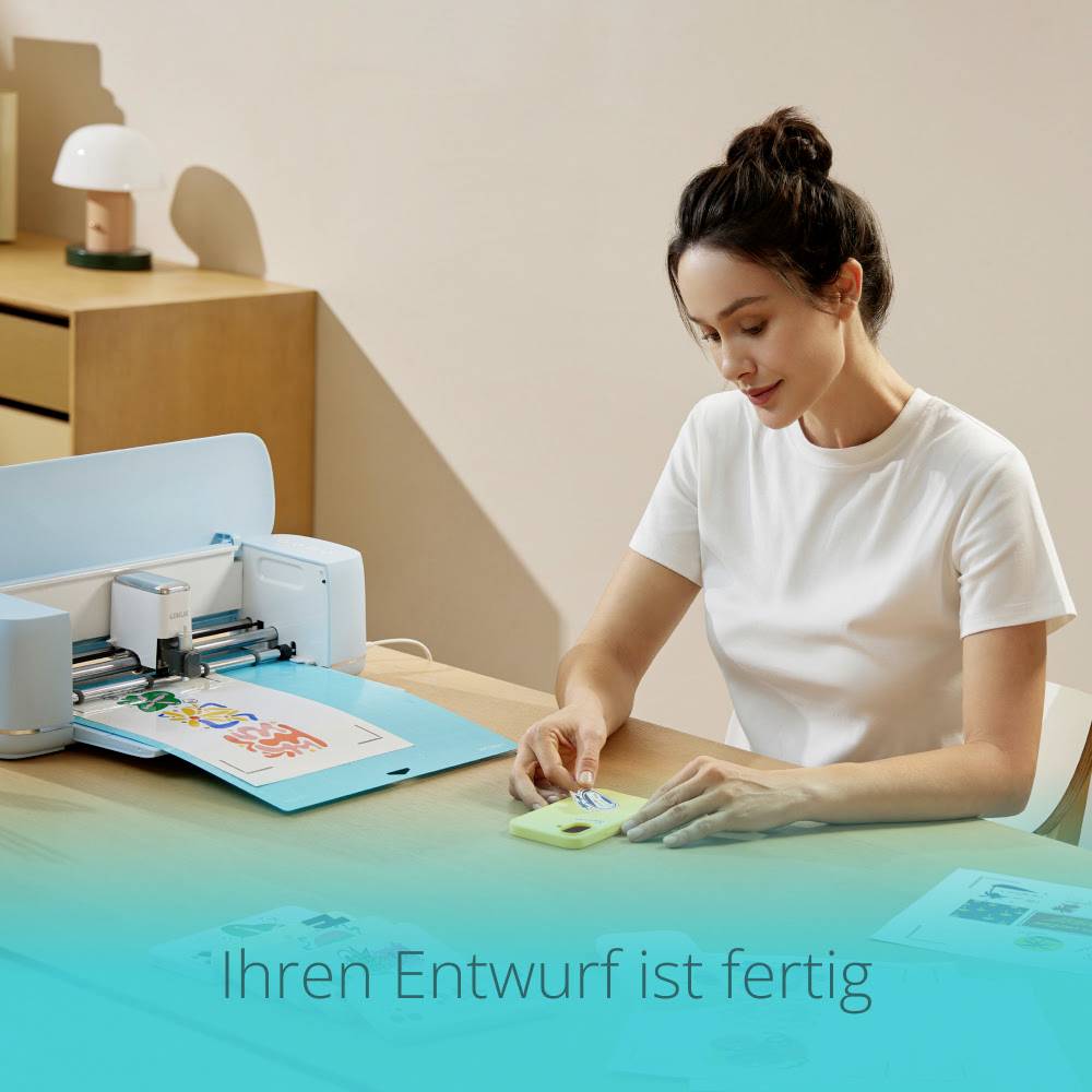A woman in a white T-shirt sits at a desk with a printer processing coloured paper. Text at the edge of the image: 'Your design is ready'.