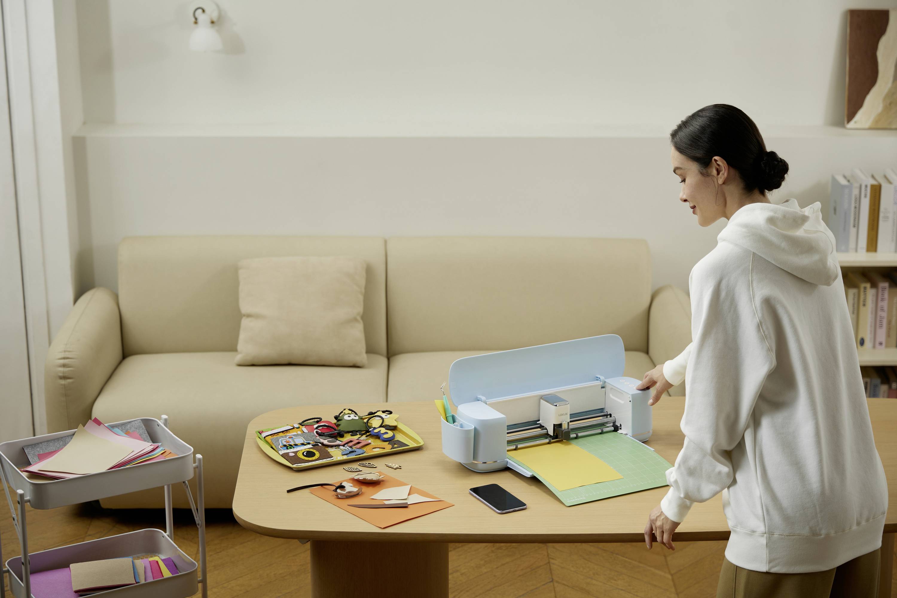 A woman is standing at a table using a craft cutting machine. The table is covered with colourful paper and tools. A sofa is visible in the background.