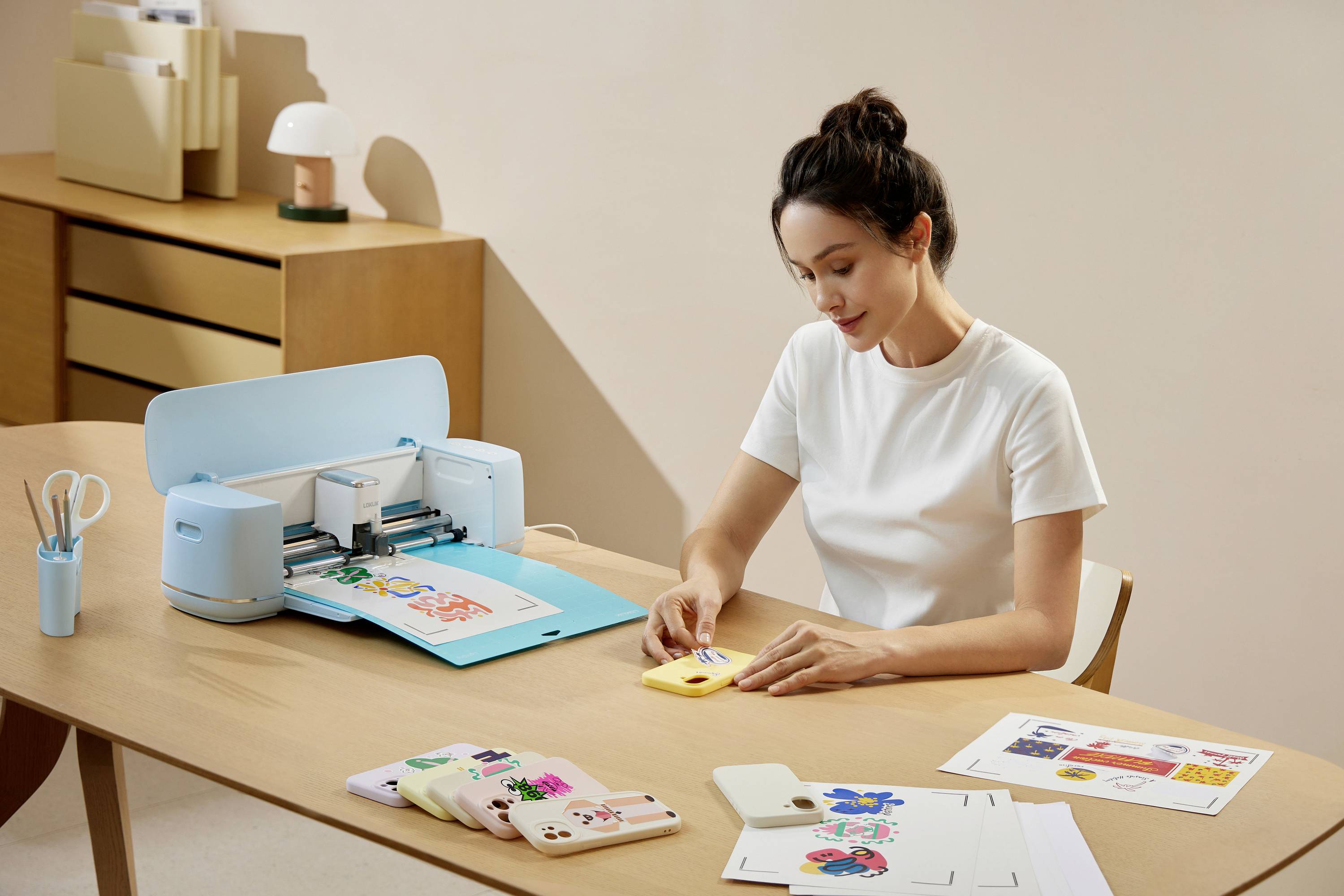 A person is sitting at a table and using a cutting machine to process colourful paper, with accessories and finished works beside them.