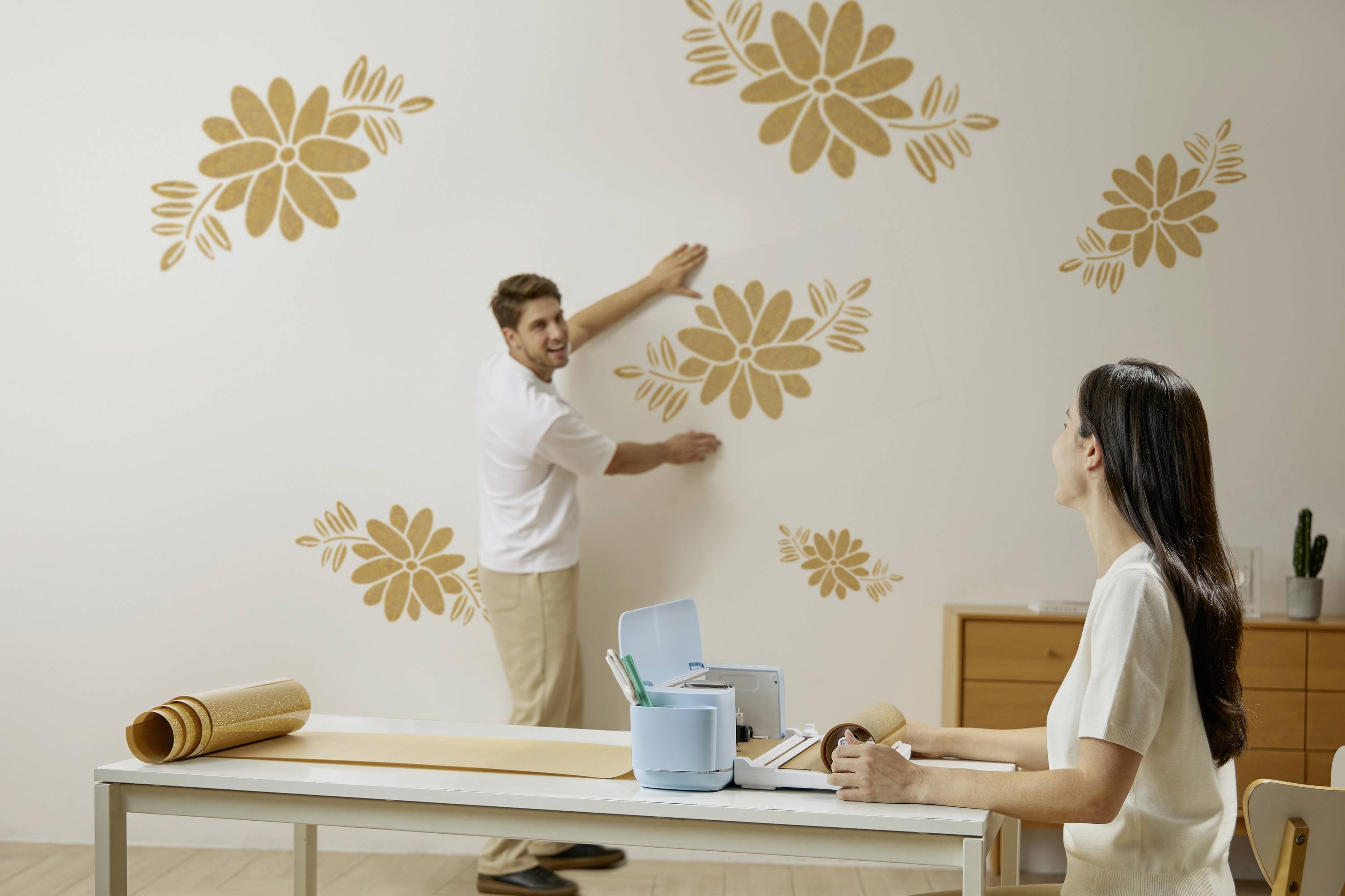 A man is sticking floral patterns onto a wall while a woman sits at a table with craft supplies, watching him.