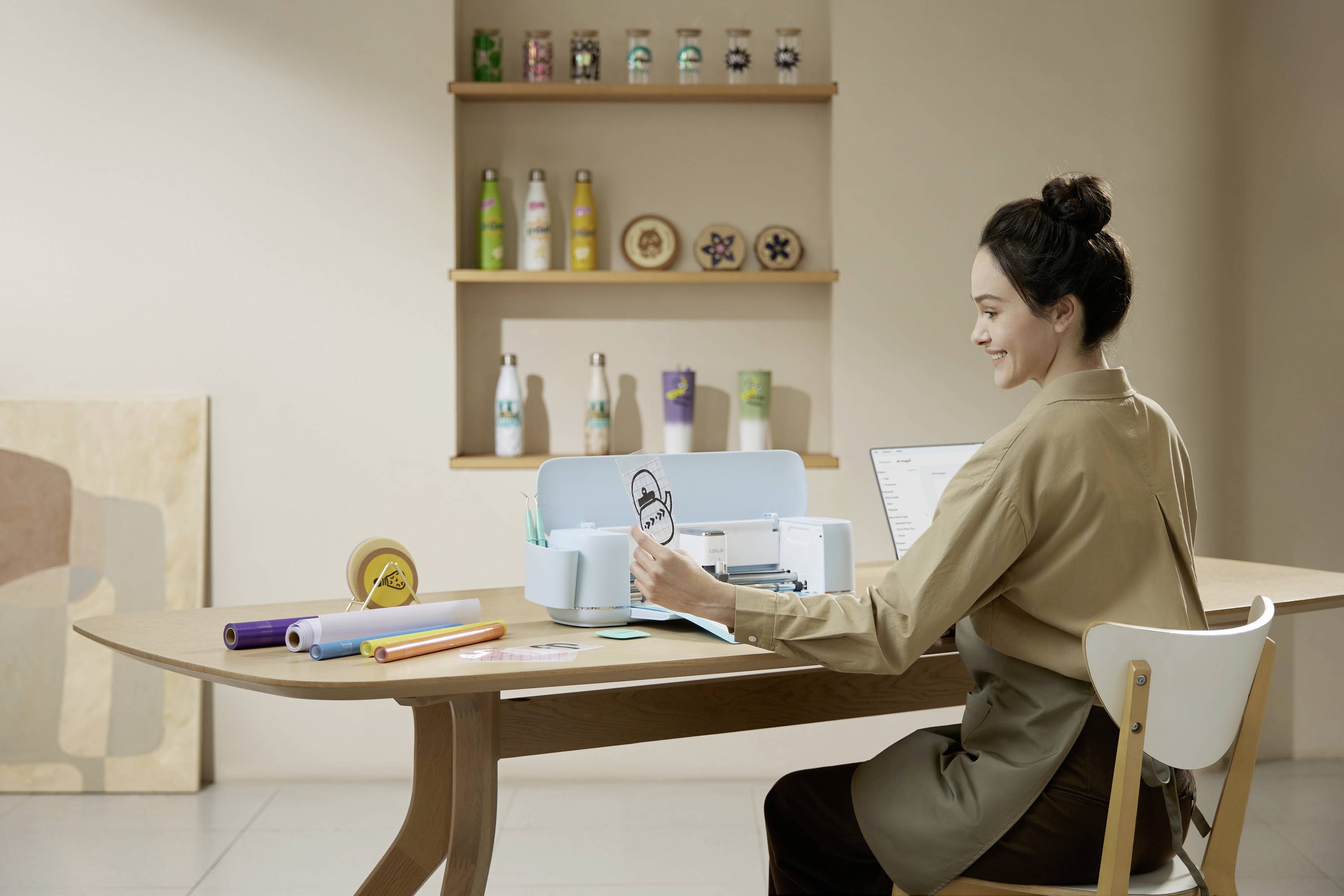 A woman is sitting at a table and using a cutting device to cut out a design from paper. Drinks and materials can be seen in the background.