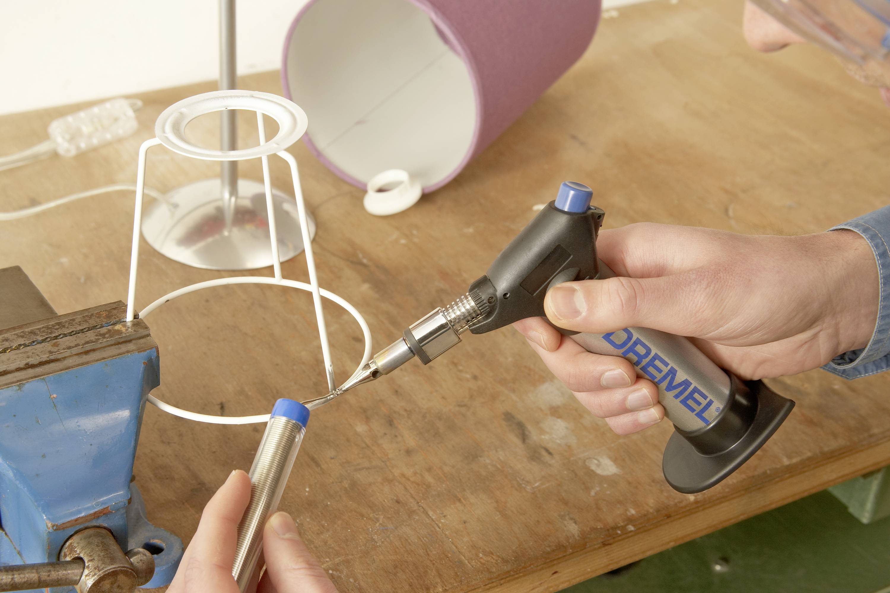 A person is working on a metal object with a blue soldering tool on a workbench. A desk lamp is lying upside down nearby.