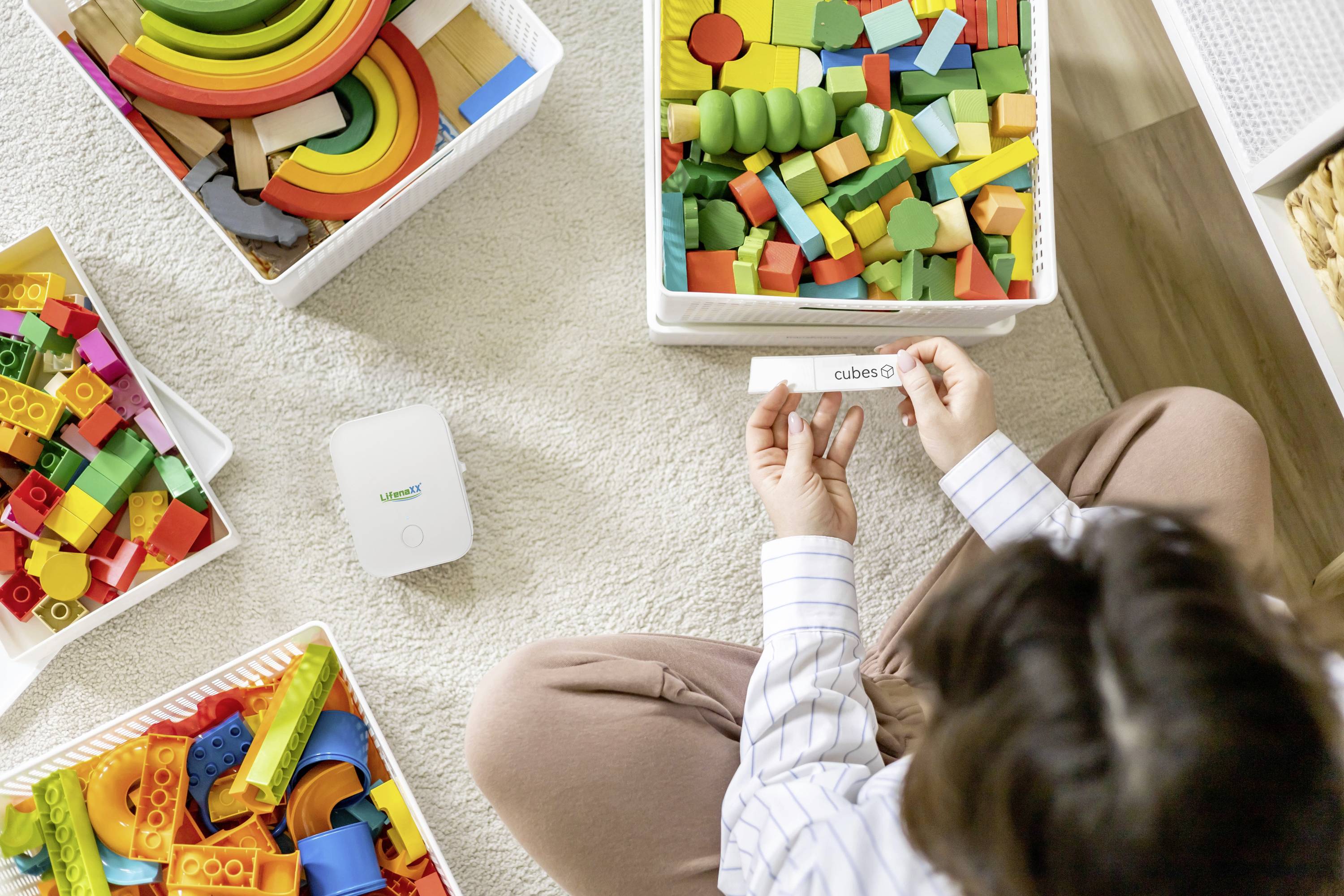 A person is sitting on the floor, sorting colourful building blocks from various containers. A label with 'cubes' is held in their hand.