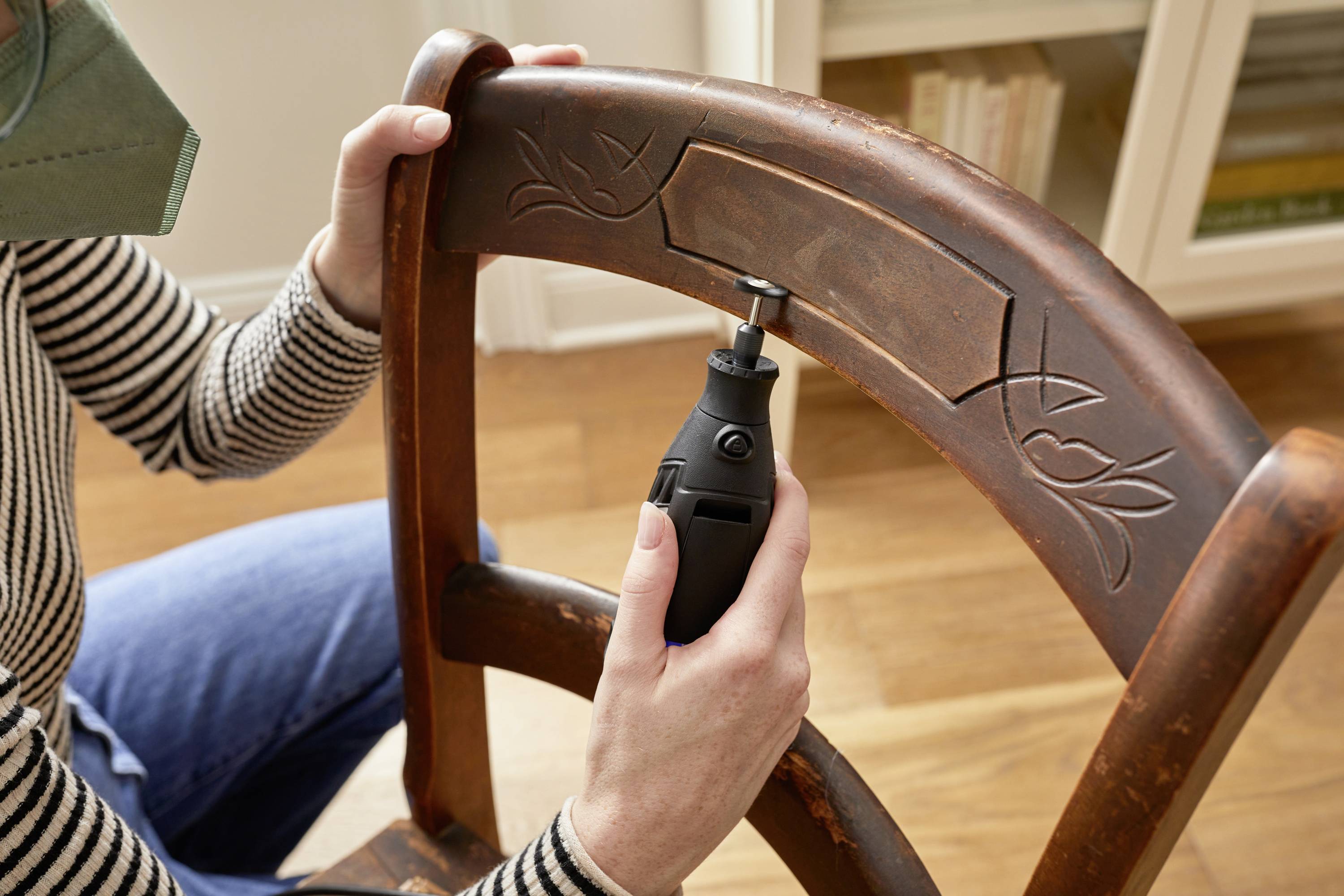 A person is repairing a wooden chair back with an electrical tool. The back features decorative engravings. A bookshelf is visible in the background.
