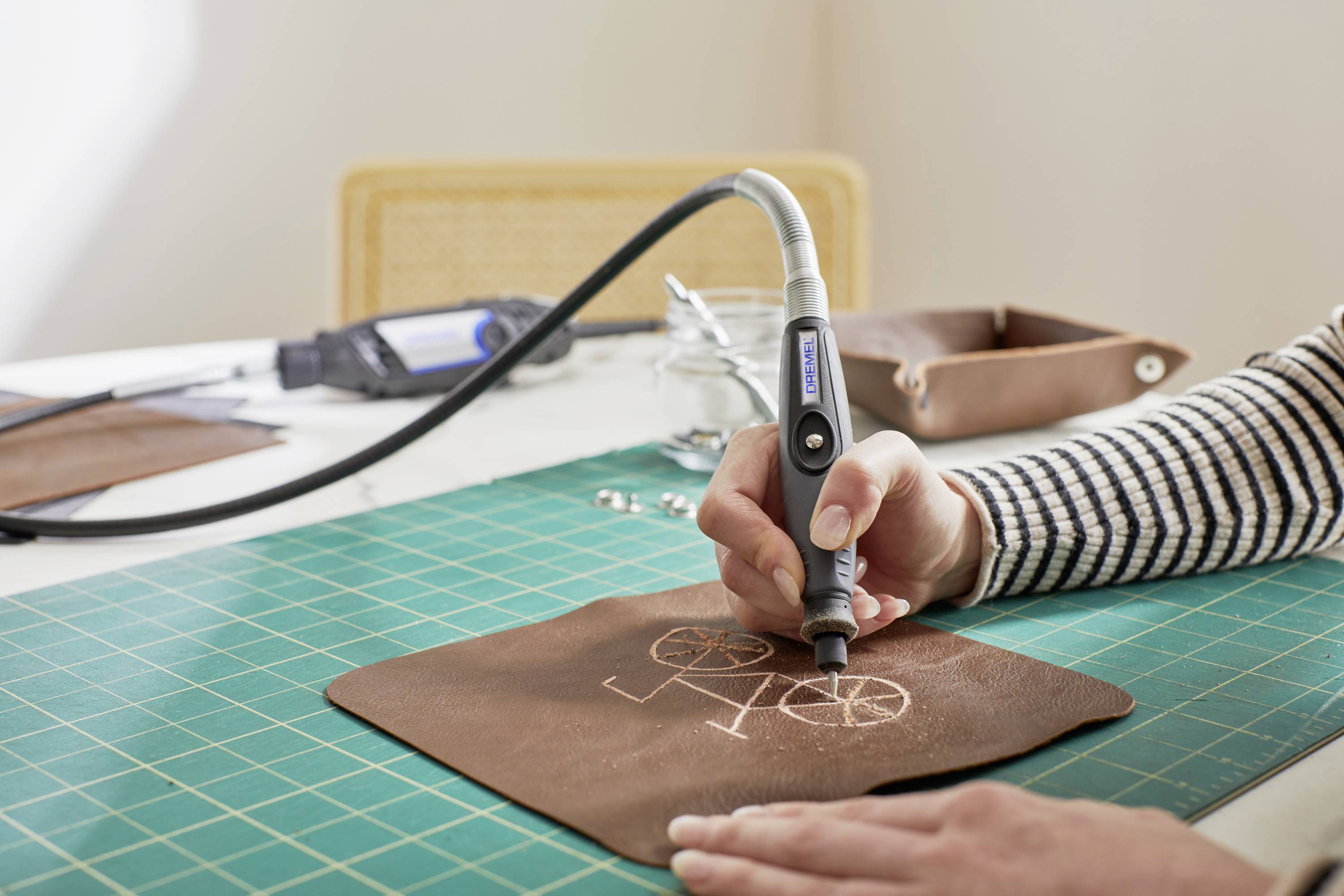 A person is engraving a bicycle motif into a piece of brown leather on a green cutting mat using an electric engraving pen.