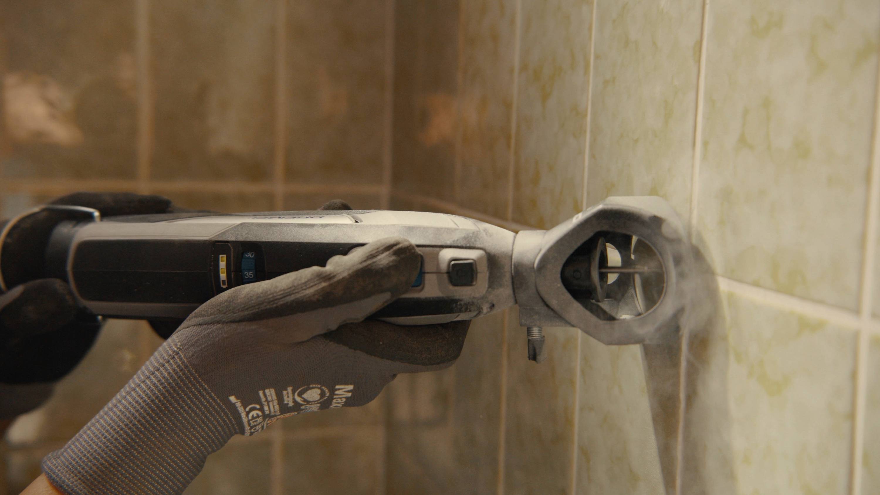 A person is using an electric multi-function tool to work on tile joints. Protective gloves are being worn.